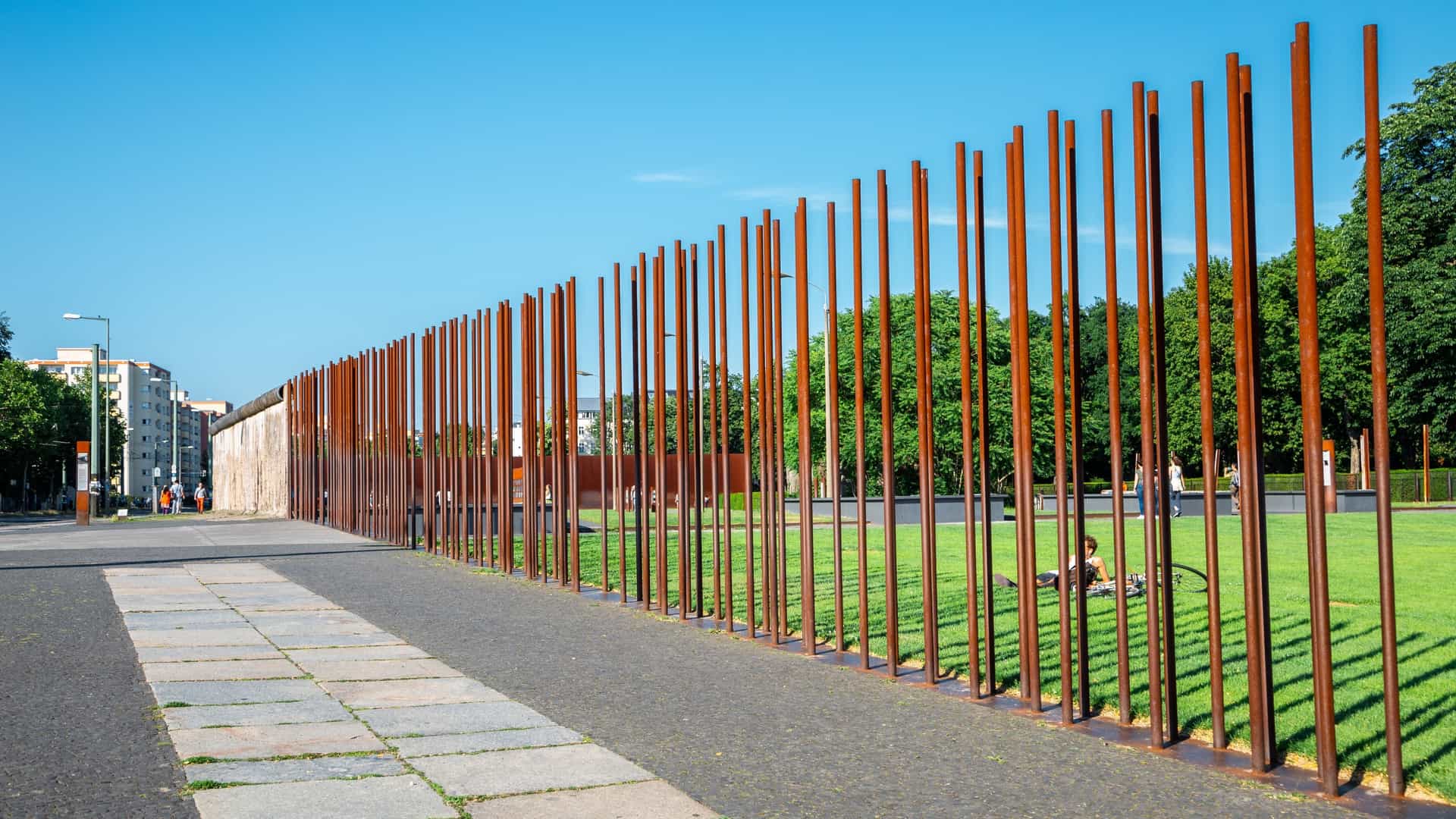 A view of the Berlin Wall Memorial (Gedenkstätte Berliner Mauer) in Germany, featuring the preserved remnants of the "death strip." In the foreground is a section of the inner wall made of concrete slabs, backed by a metal lattice fence and the watchtower. The scene is marked by the contrasting elements of the oppressive Cold War border infrastructure against a backdrop of green grass and contemporary Berlin buildings.