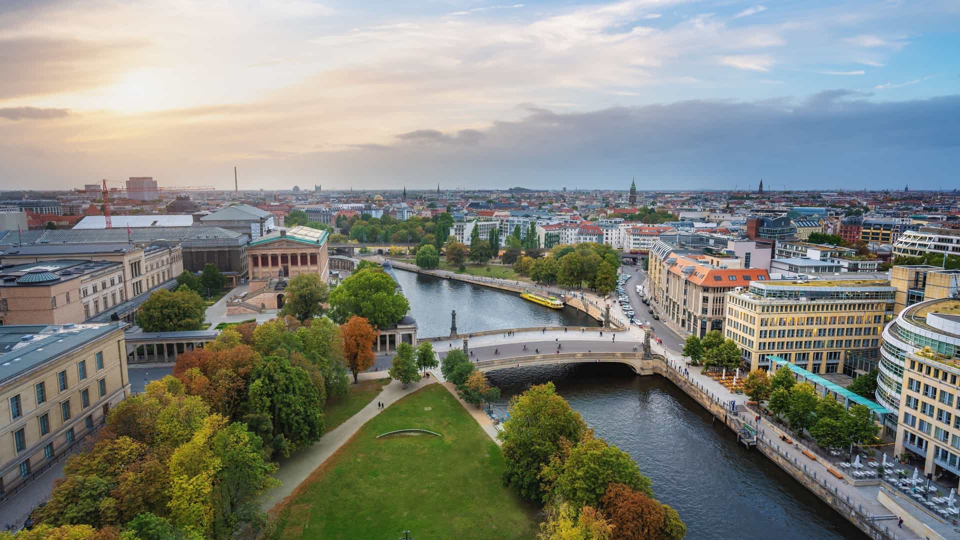 An aerial view of Berlin's Museum Island on a sunny day, with the Spree River flowing around it. The historic Berlin Cathedral and the domed Bode Museum are prominent among the buildings on the island.