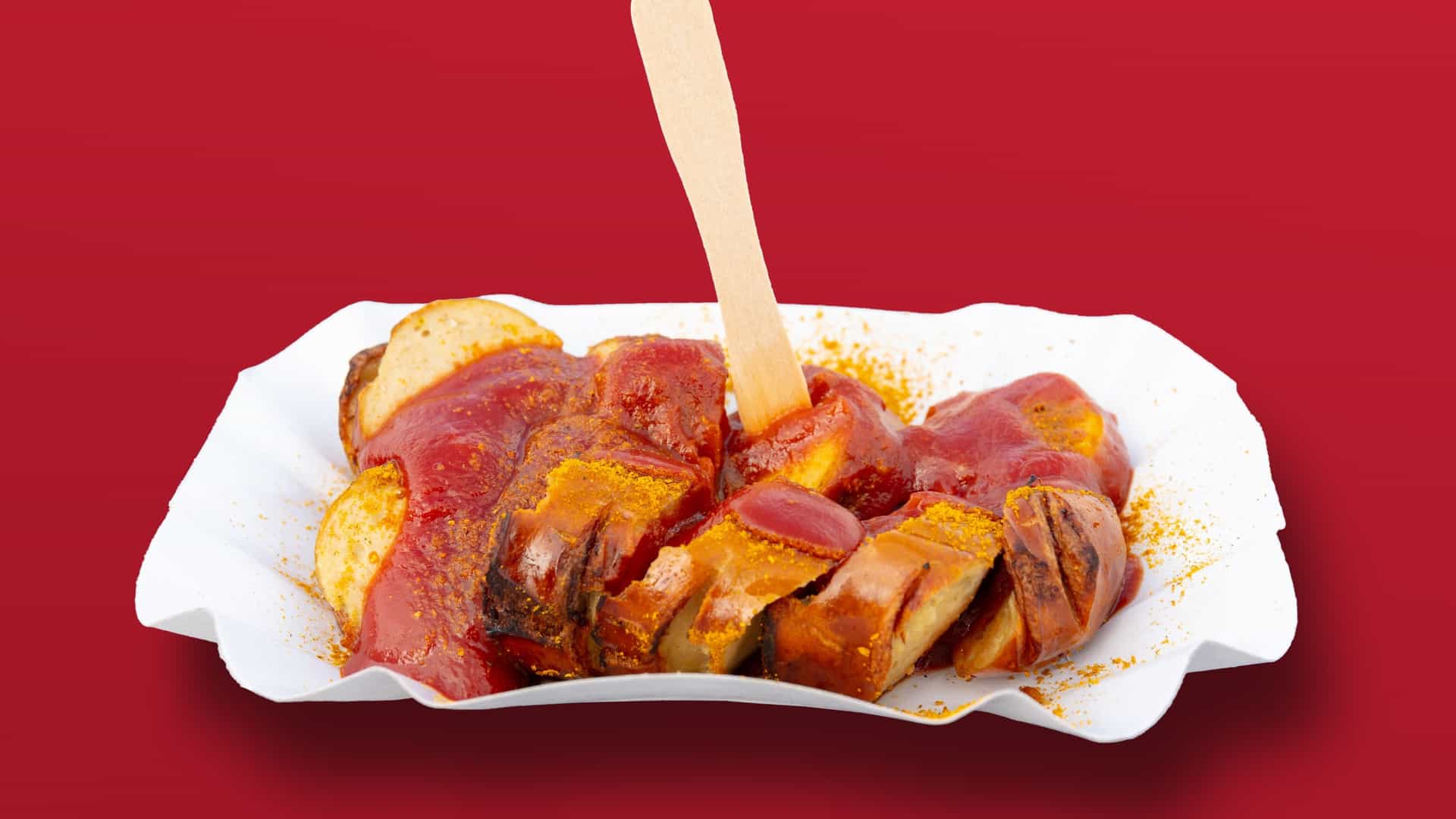 A close up, high angle view of a serving of Currywurst, a popular German street food. Sliced grilled sausage (Wurst) is completely smothered in a thick, reddish brown curry ketchup sauce and dusted generously with bright yellow curry powder. The dish is presented on a white plate with a side of golden brown French fries (Pommes Frites) and a small dollop of white mayonnaise. The meal is set on a rustic wooden table.