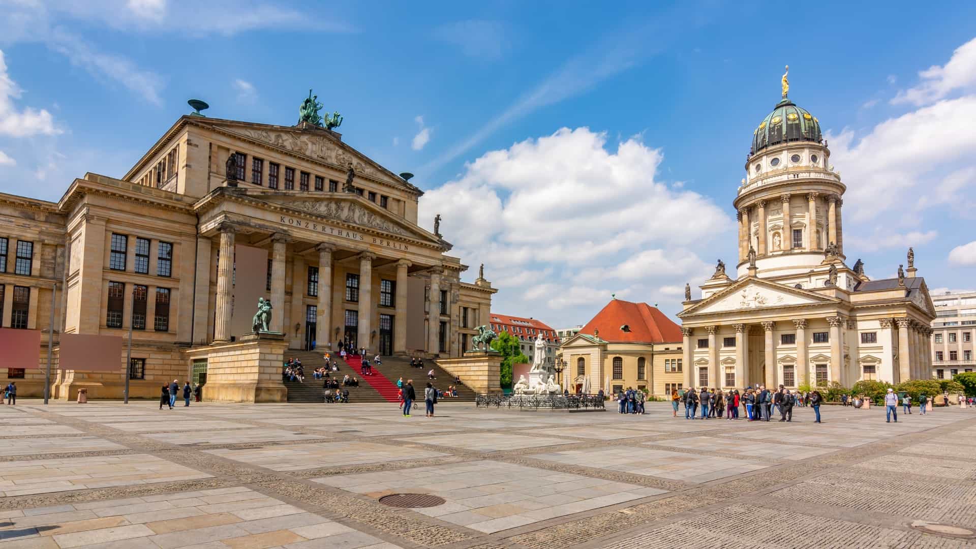 The magnificent Gendarmenmarkt square in Berlin, Germany, framed by two of its neoclassical architectural landmarks. The image is taken from the steps of the Konzerthaus Berlin (Concert Hall) in the foreground, looking toward the distant Französischer Dom (French Cathedral). The cathedral's distinctive domed tower rises above the square, which is currently undergoing renovation and surrounded by high green construction fences. The sky above is bright blue with a scattering of white clouds.