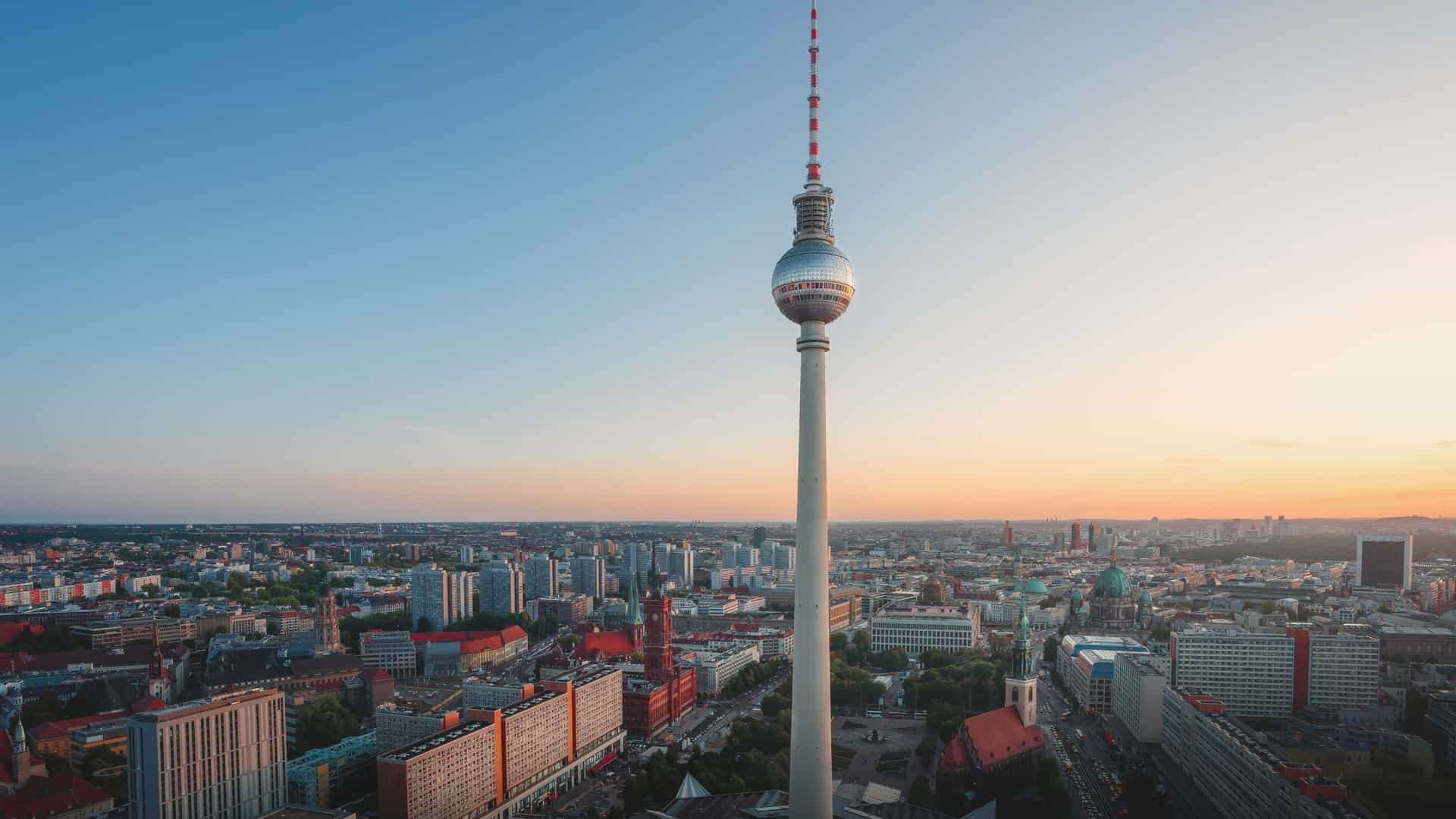 An aerial view of the Berlin skyline with the iconic Berlin Television Tower, or Fernsehturm, at its center. The tower's slender spire and large, spherical observation deck rise above the surrounding buildings, showcasing its status as a major landmark in the city.