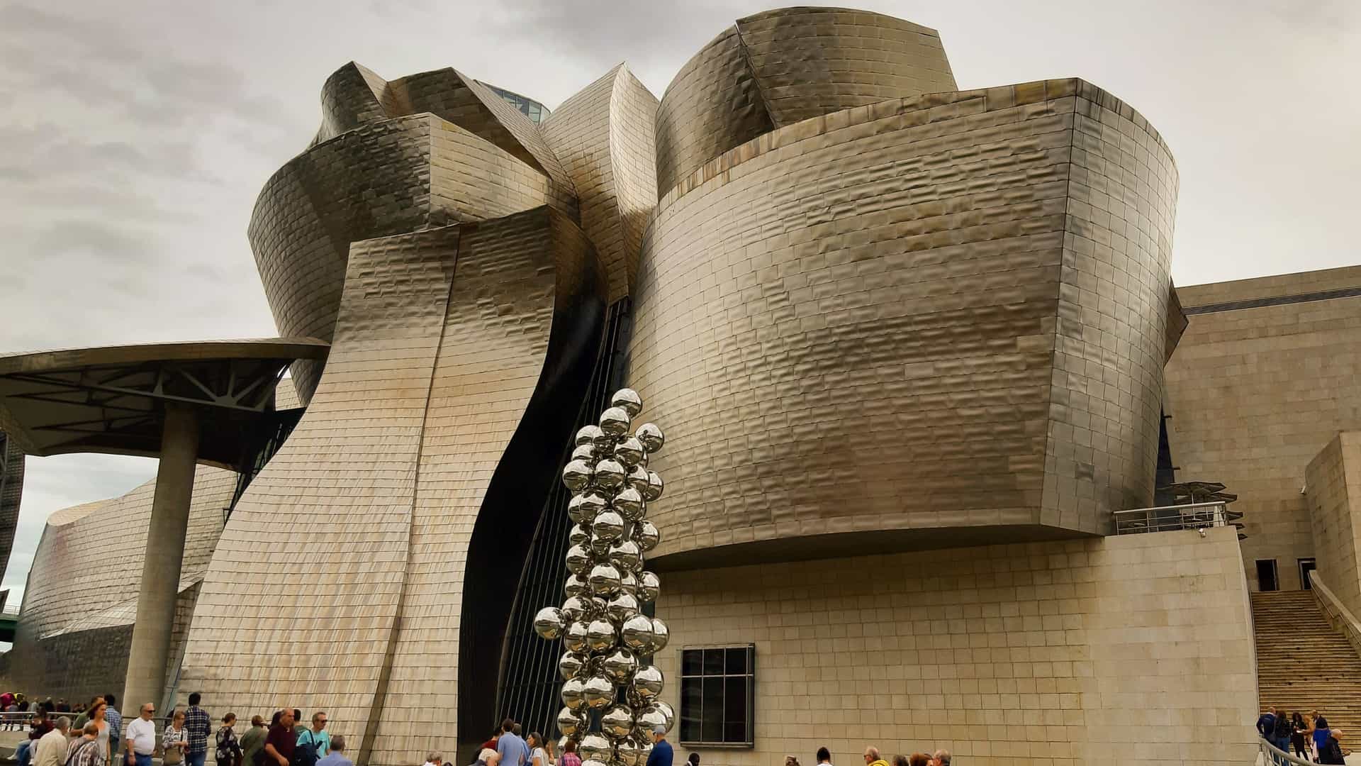 A bright, panoramic view of the Guggenheim Museum Bilbao in the Basque Country, Spain. The deconstructivist building, designed by Frank Gehry, features its signature undulating, titanium clad curves reflecting the light. In the foreground, the sculpture Puppy by Jeff Koons, a giant topiary of a West Highland Terrier, stands covered in colorful flowers. The museum is set beside the Nervión River, with modern and traditional city buildings visible in the background under a blue sky.