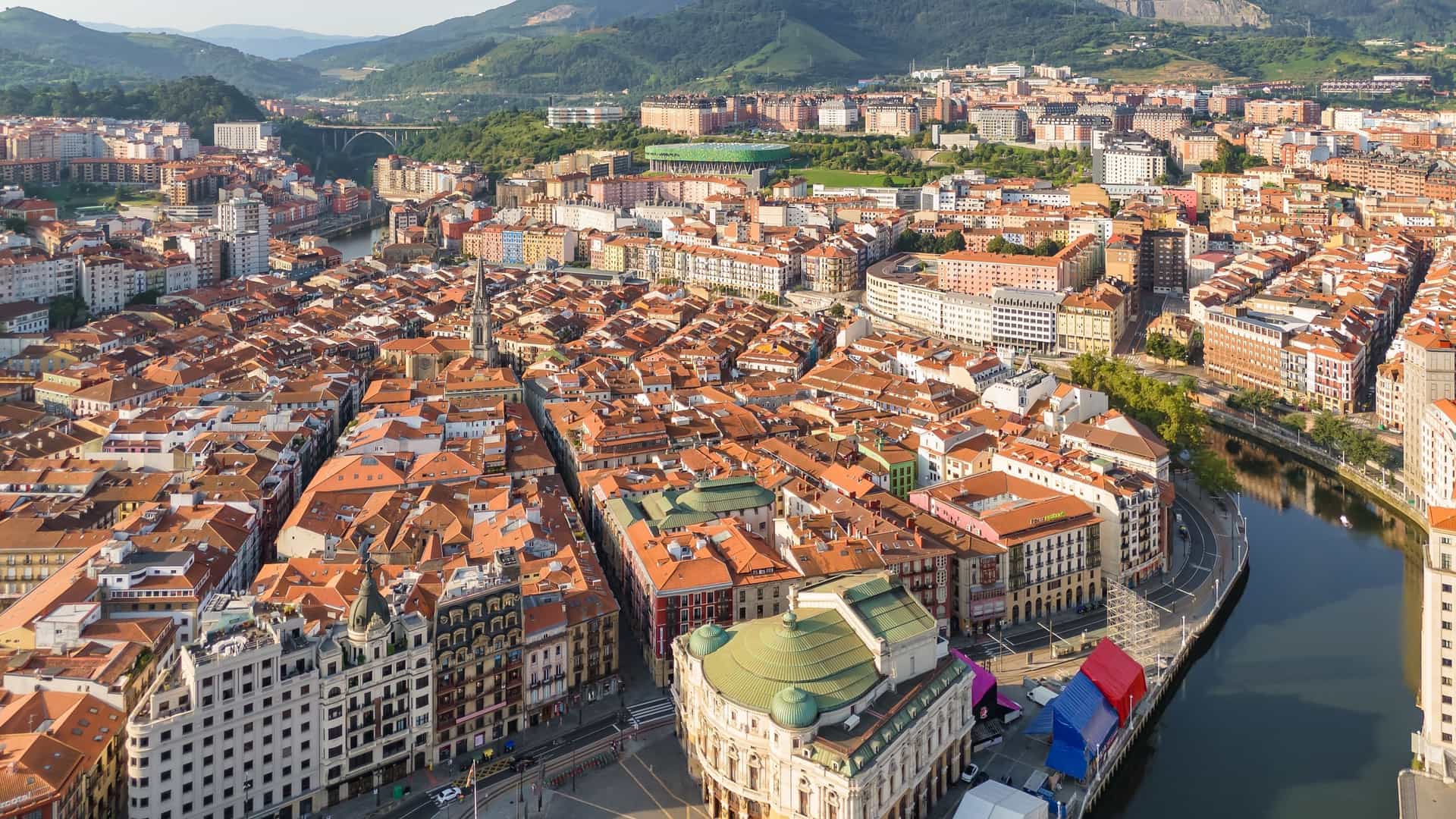 An aerial, bird's eye view of the dense, historic Old Town (Casco Viejo) of Bilbao, Basque Country, Spain. The image features a maze of tightly packed buildings with characteristic red tiled rooftops and the historic grid of the seven original streets.