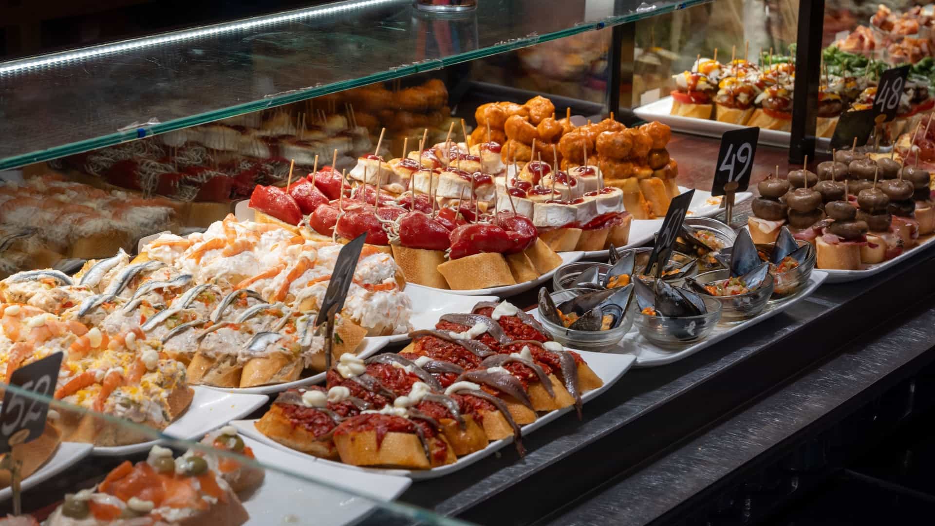 A close up view of a bar counter showcasing a vibrant, diverse array of traditional Basque pintxos (or pinchos), which are small snacks typically skewered with a toothpick onto a piece of bread. The selection includes toppings like seafood, fish, eggs, and sweet green peppers.