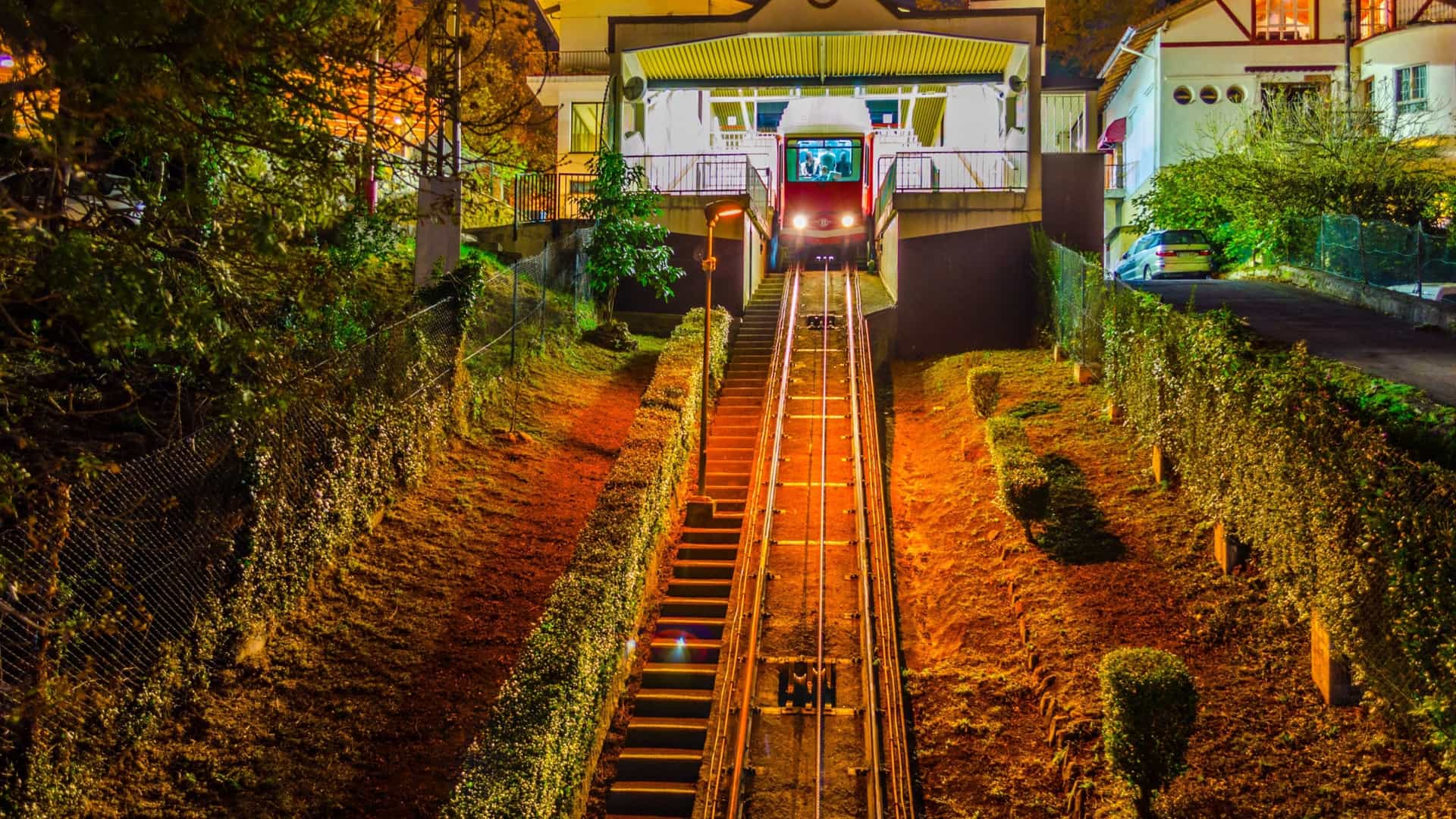 A night view of the Artxanda Funicular station in Bilbao, Spain. The image features the brightly illuminated funicular cabin and its tracks leading up a steep slope, set against the dark background of the mountain or city night sky.