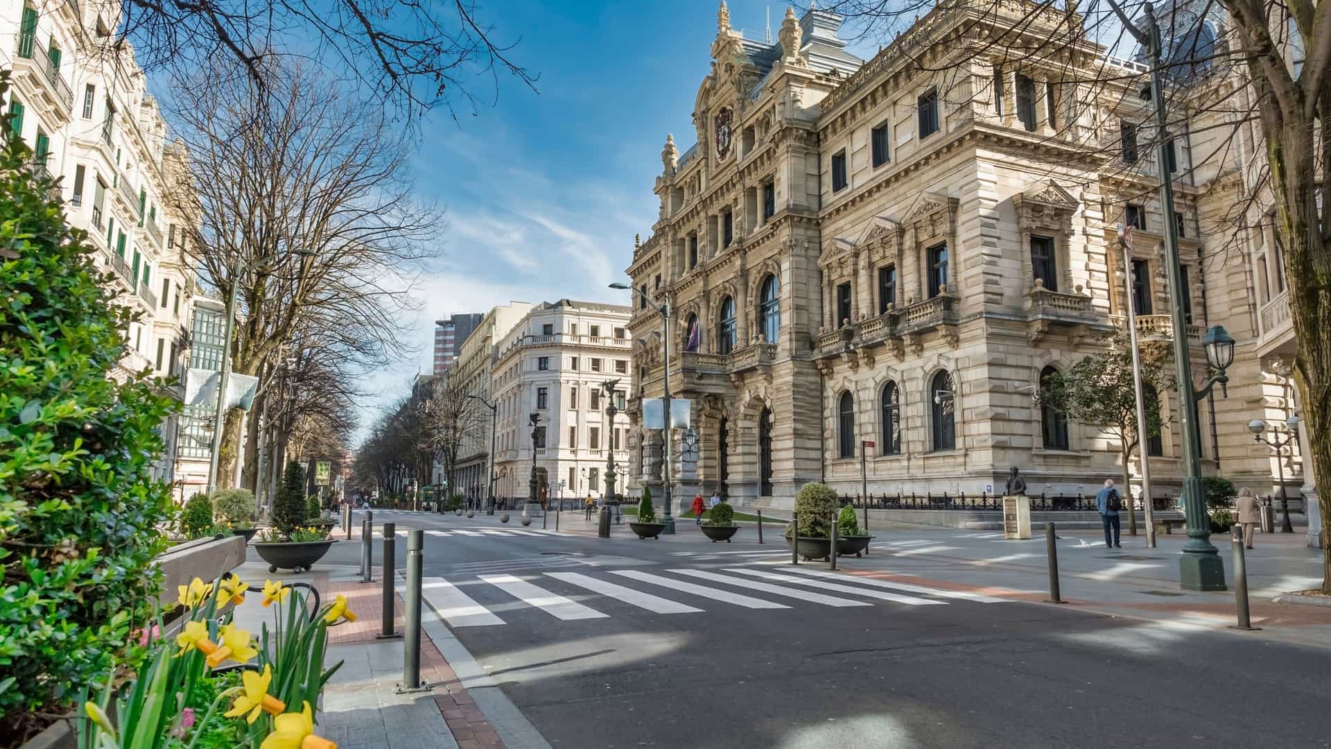 A wide angle view of an elegant street in the Ensanche district of Bilbao, Spain. On the right is a grand, neoclassical style building with ornate stone work and arched windows. The street features a wide pedestrian area with a crosswalk, street lamps, and planters containing yellow and orange daffodils in the foreground. Bare branched trees line the sidewalks under a clear blue sky.