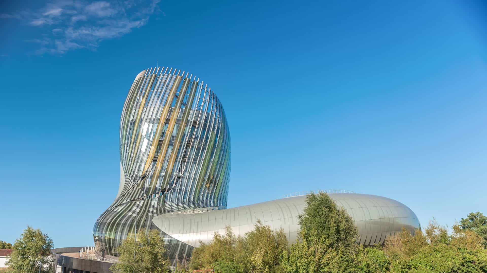 The modern, futuristic exterior of La Cité du Vin (The City of Wine) museum in Bordeaux, France. The building has an iconic, bulbous, curved form that is designed to evoke wine swirling in a glass or the eddies of the nearby Garonne River. Its facade is a patchwork of reflective, perforated, iridescent aluminum and silk screened glass panels, shimmering in a golden hue against a partly cloudy sky. The structure is set along the waterfront, with a plaza and light colored pavement visible in the foreground.