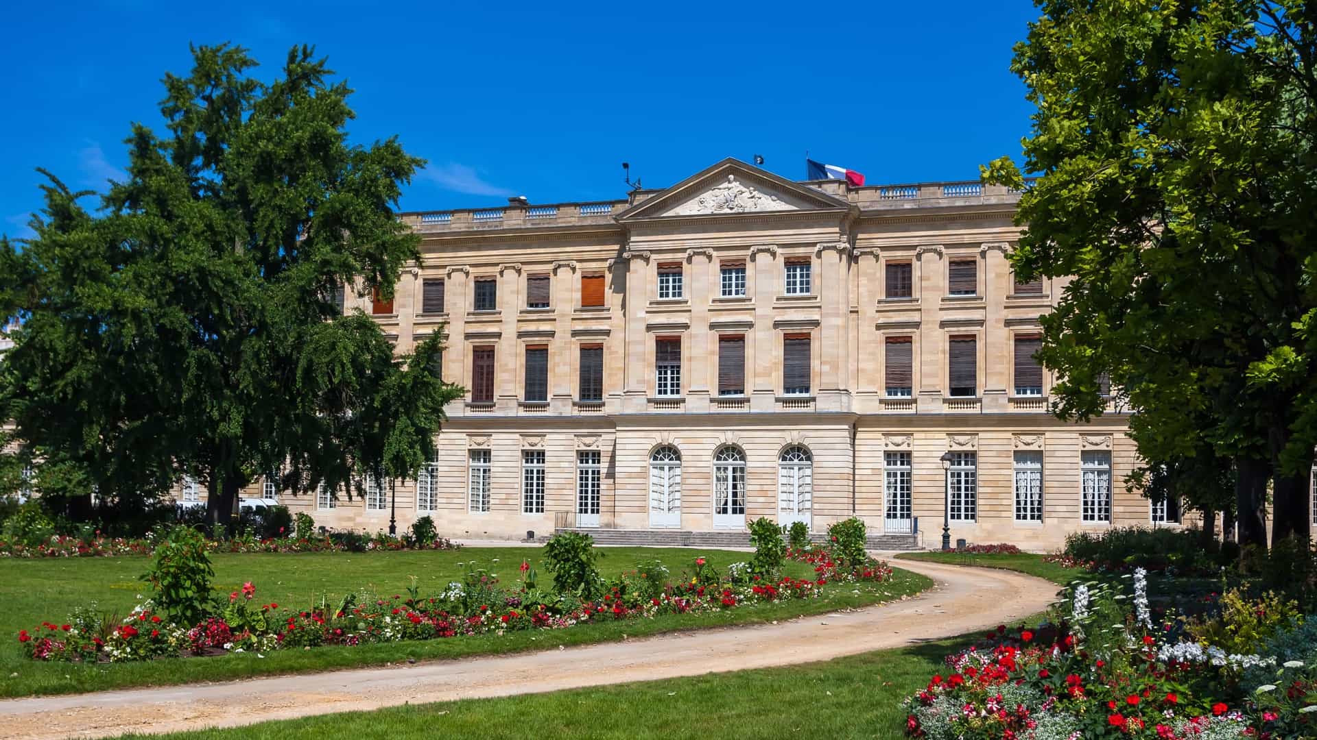 The facade of the Musée des Beaux Arts de Bordeaux (Fine Arts Museum of Bordeaux), which is part of a complex located in the gardens of the city hall. The building features classical, stately architecture, characterized by light colored stone. The photograph shows a section of the museum's exterior, which has large, arched windows and is surrounded by manicured green space and trees. The classical details of the museum's architecture are prominent under a bright sky.