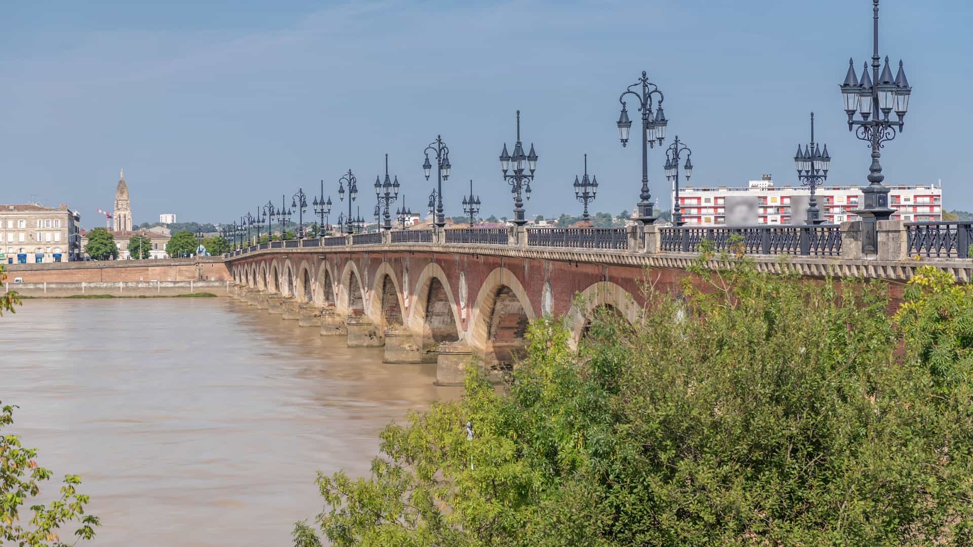 A serene long exposure view of the Pont de Pierre (Stone Bridge) spanning the Garonne River in Bordeaux, France. The bridge's historic stone arches and antique lampposts are illuminated, creating a soft glow that reflects on the calm water, with streaks of light from passing vehicles visible on the roadway. The sky is in the deep blue of twilight.