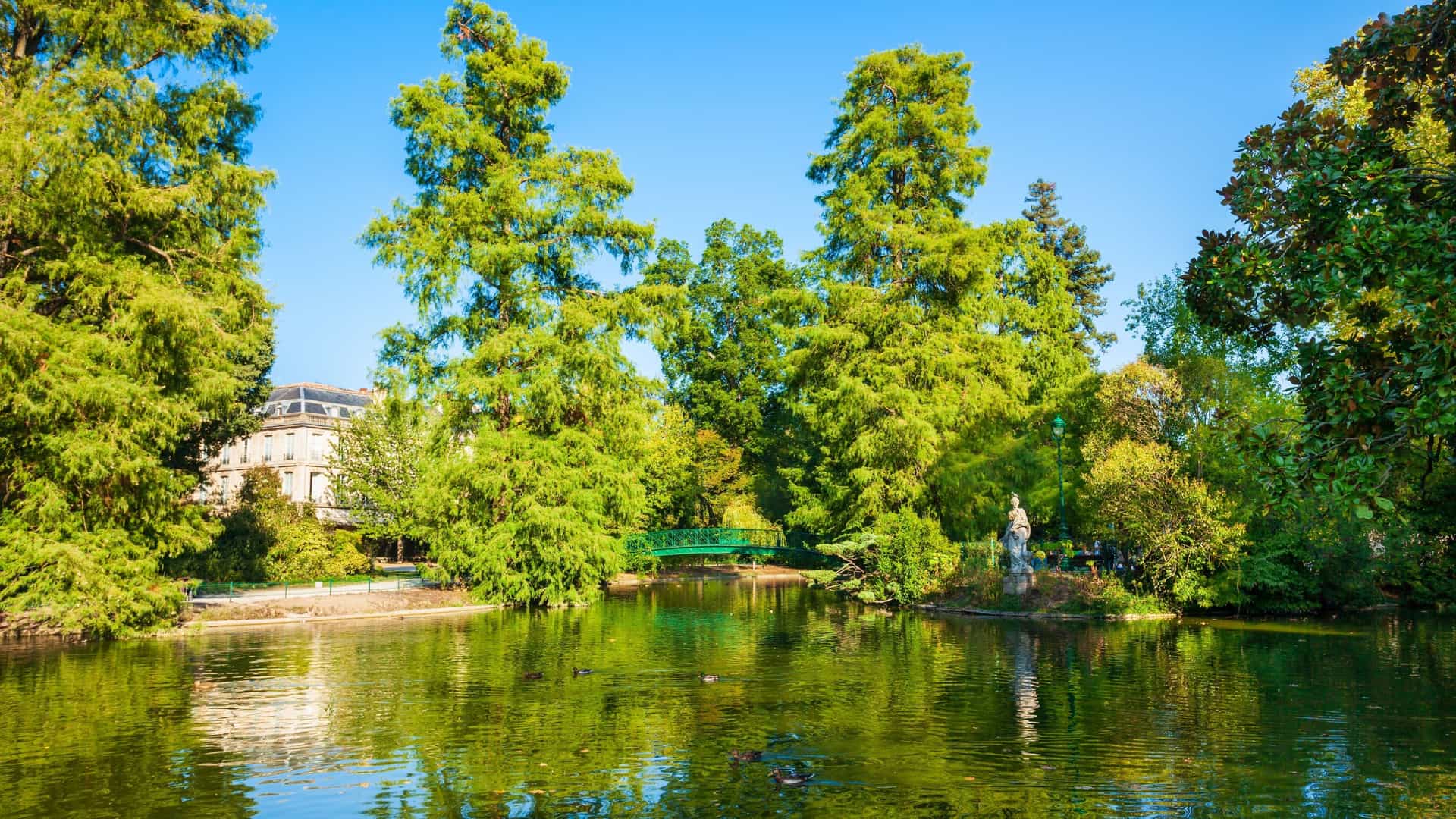 A scenic view of the Jardin Public in Bordeaux, France. The image shows a serene central pond with a small, arched bridge and lush greenery along the banks. In the background, elegant, 18th century stone buildings frame the garden, highlighting its status as a historical and tranquil escape in the city center.
