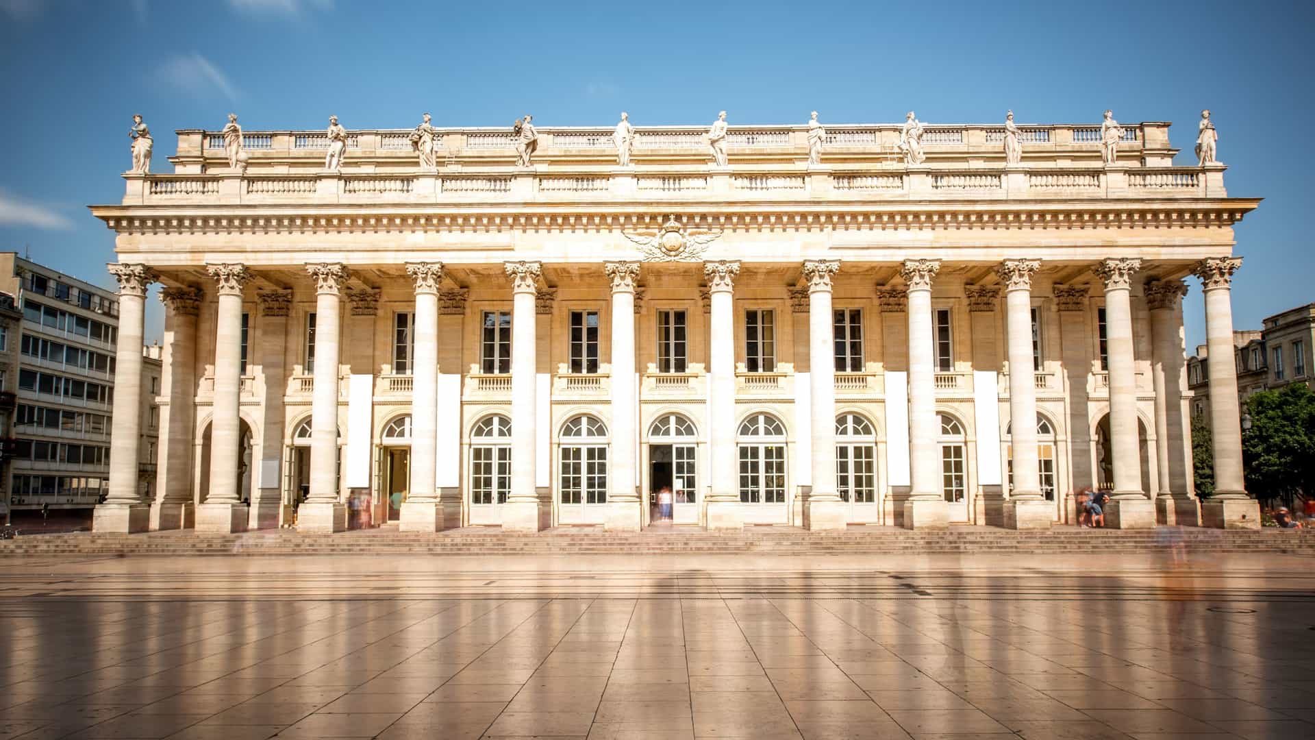 A long exposure view of the Grand Théâtre de Bordeaux, a neoclassical opera house in France. The facade features a colonnade of twelve Corinthian columns topped with a balustrade adorned with statues. The long exposure technique blurs the movement of people and clouds, giving the scene a soft, ethereal quality. The building is a prominent landmark on the Place de la Comédie.