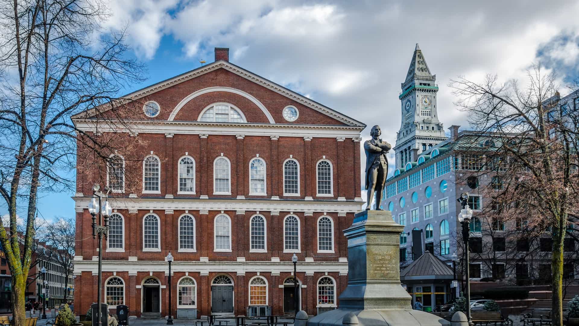 The historic Faneuil Hall, a red brick, three story building, stands within the wider Faneuil Hall Marketplace area in Boston, Massachusetts. A bronze statue of Samuel Adams on a stone pedestal is visible in the foreground, with the Customs House Tower clock in the background.