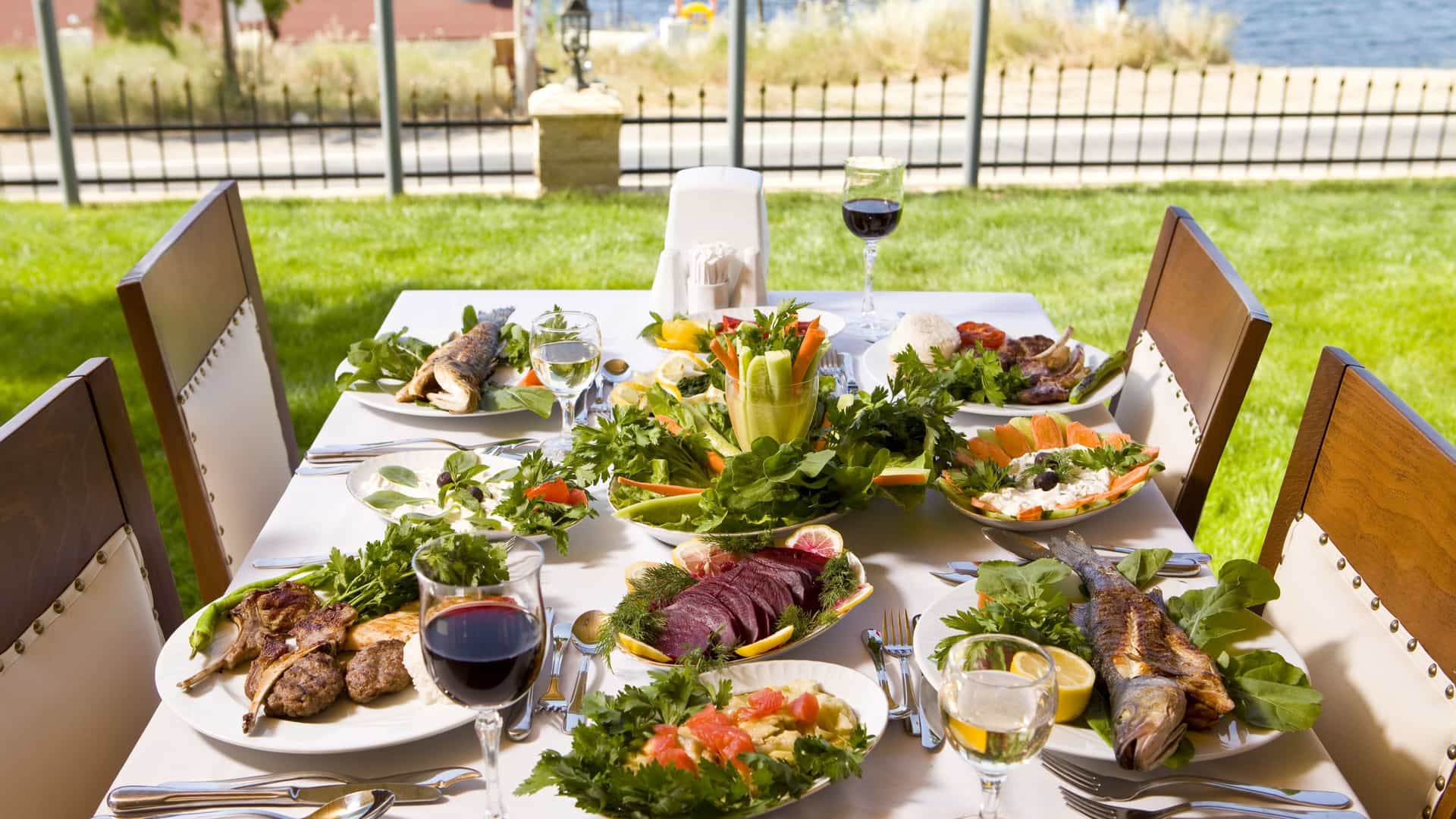 A lavish outdoor dining experience, potentially representing Dining in the Seaport District in Boston, featuring a table set with multiple plates of grilled fish, meat, and fresh Mediterranean style salads on a sunny grassy lawn overlooking the water.