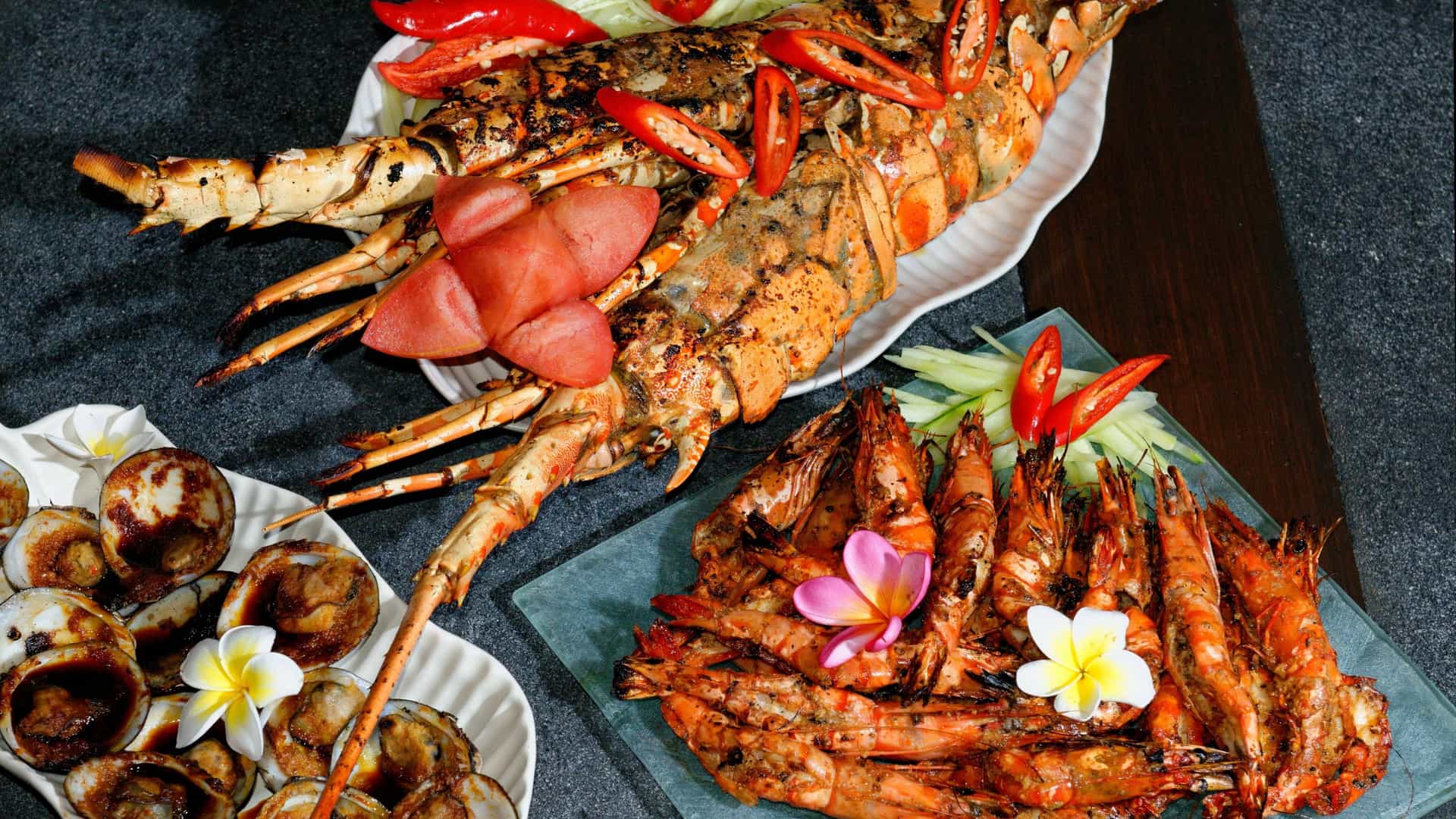 A close up view of a grilled seafood platter featuring a split, grilled lobster and several jumbo prawns (or shrimp) in their shells. The platter is typically garnished with fresh herbs and lemon wedges.