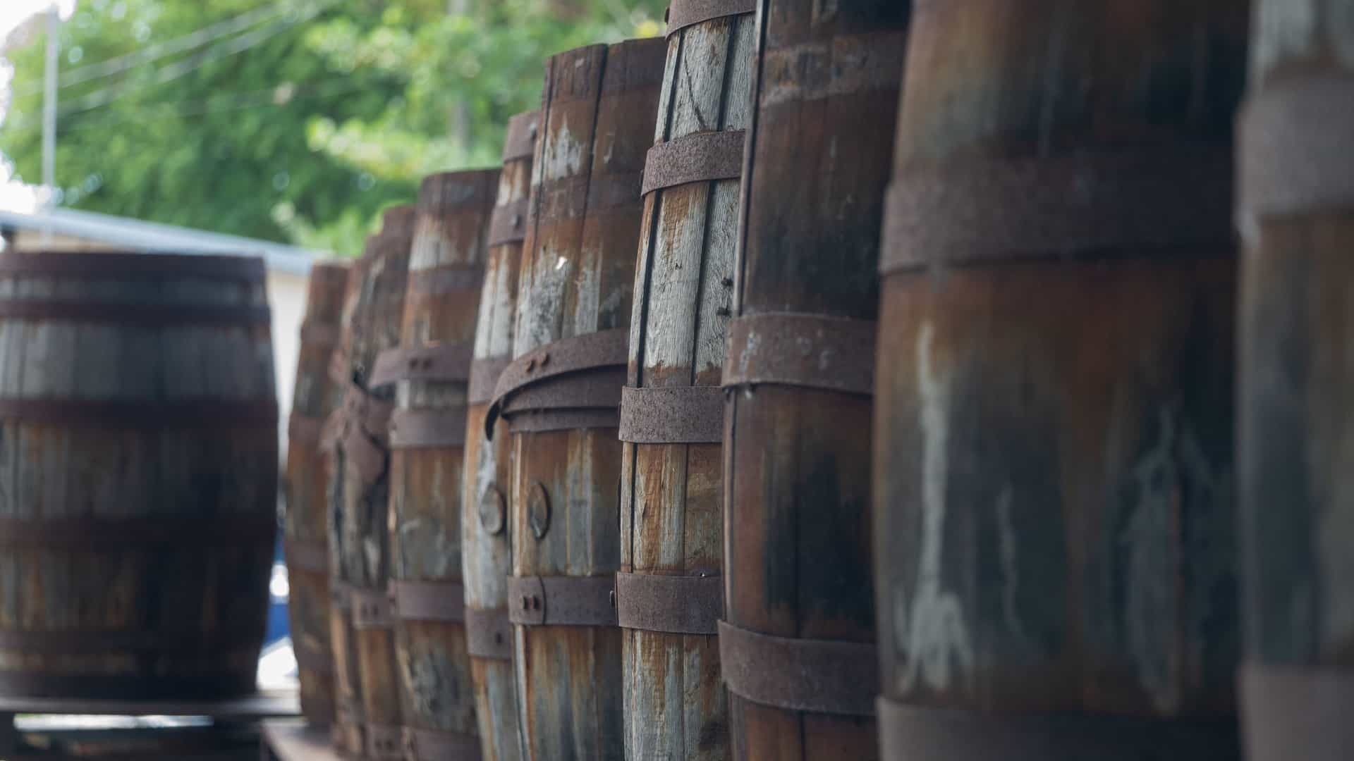 A close up of a large stack of old wooden rum barrels, likely from the Mount Gay Rum Distillery, in a dimly lit aging cellar in Barbados. The wooden casks have a rich, dark color and are held together by rusty metal bands, conveying the traditional process of aging rum.