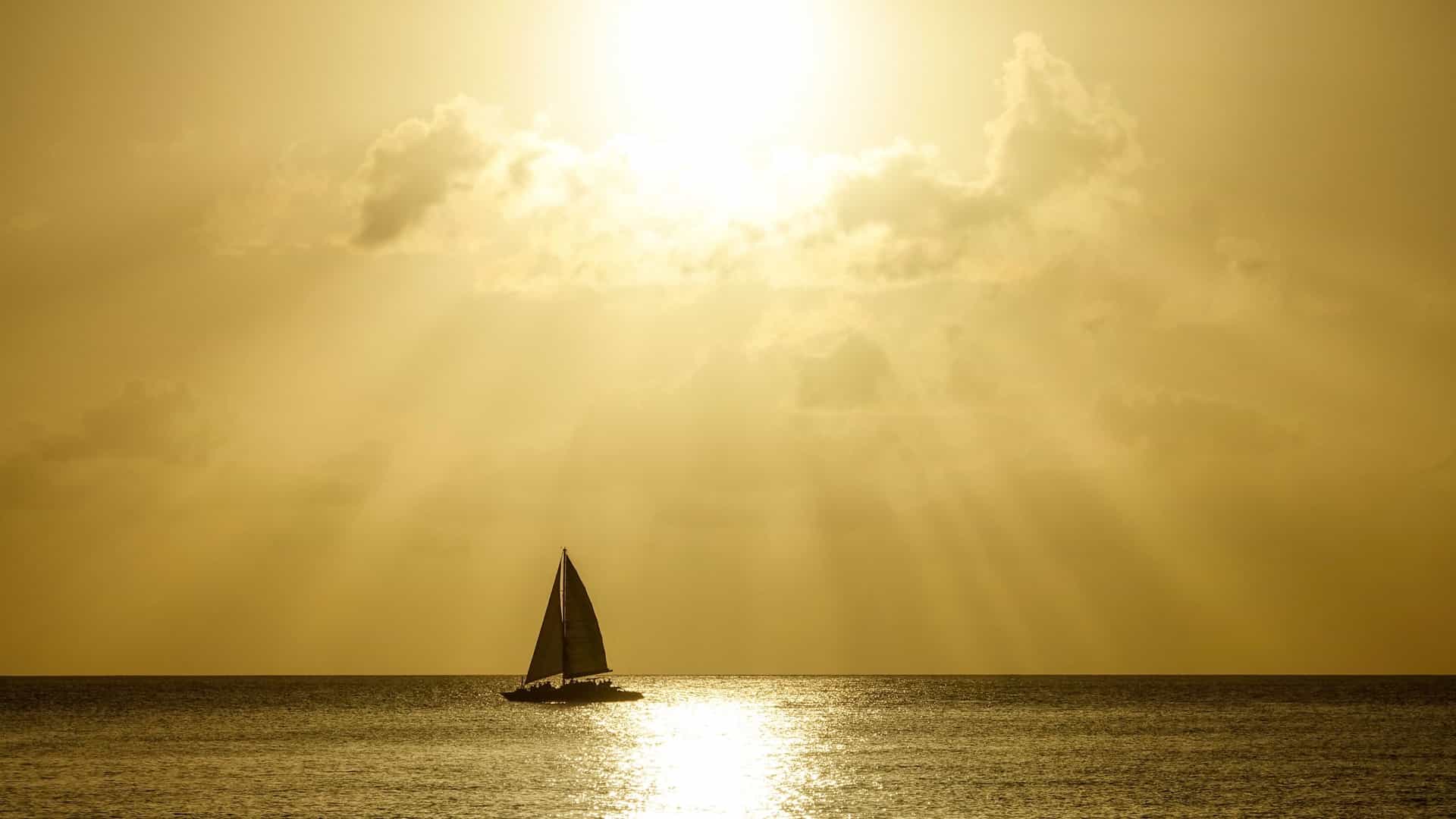 A serene image of a sailboat as a silhouette on a tranquil sea at sunset. The sky is a blend of warm colors, including shades of orange and pink, which are reflected on the calm water's surface. The boat's dark silhouette and raised sails stand out against the glowing sky, conveying a sense of peace and solitude.
