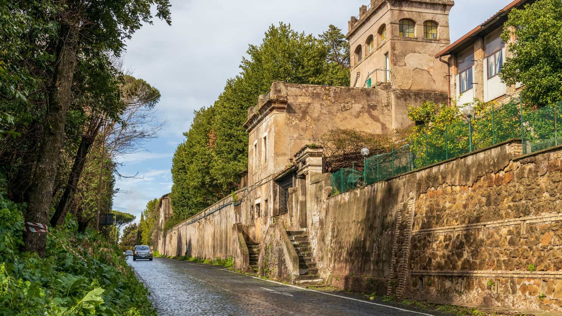 A scenic view of the Via Appia Antica (Old Appian Way) near Rome, Italy, featuring the ancient Roman road paved with large stones, lined by pine trees and historical ruins.