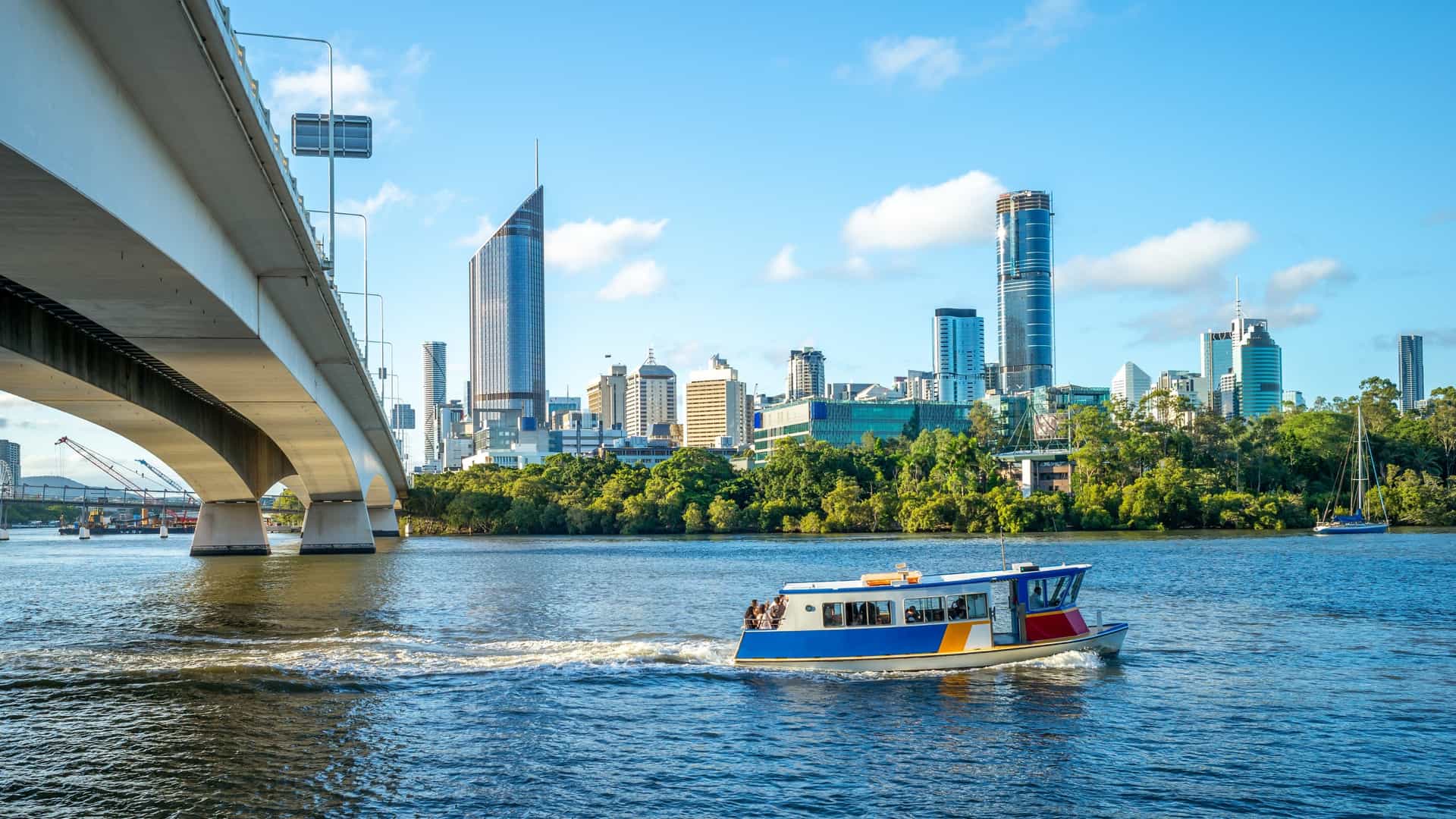 A modern ferry, likely a CityCat, cruises on the Brisbane River during the day. The ferry is heading towards the city center, with a view of the skyline and some of its prominent buildings visible in the background under a blue sky. The image captures a typical scene of public transport on the river.