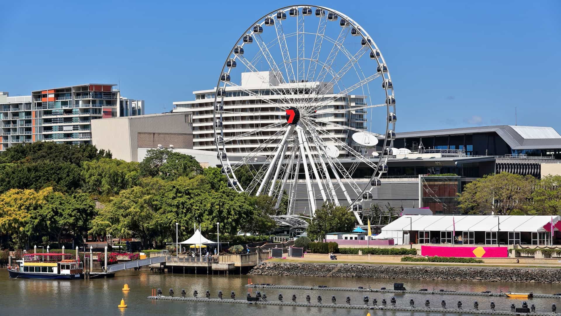The Wheel of Brisbane, a large white Ferris wheel with a red hub, stands prominently against a bright blue sky on the South Bank of the Brisbane River, Australia. The foreground shows the river with black floating pontoons, while a backdrop of modern high rise buildings and lush green trees completes the city scene.