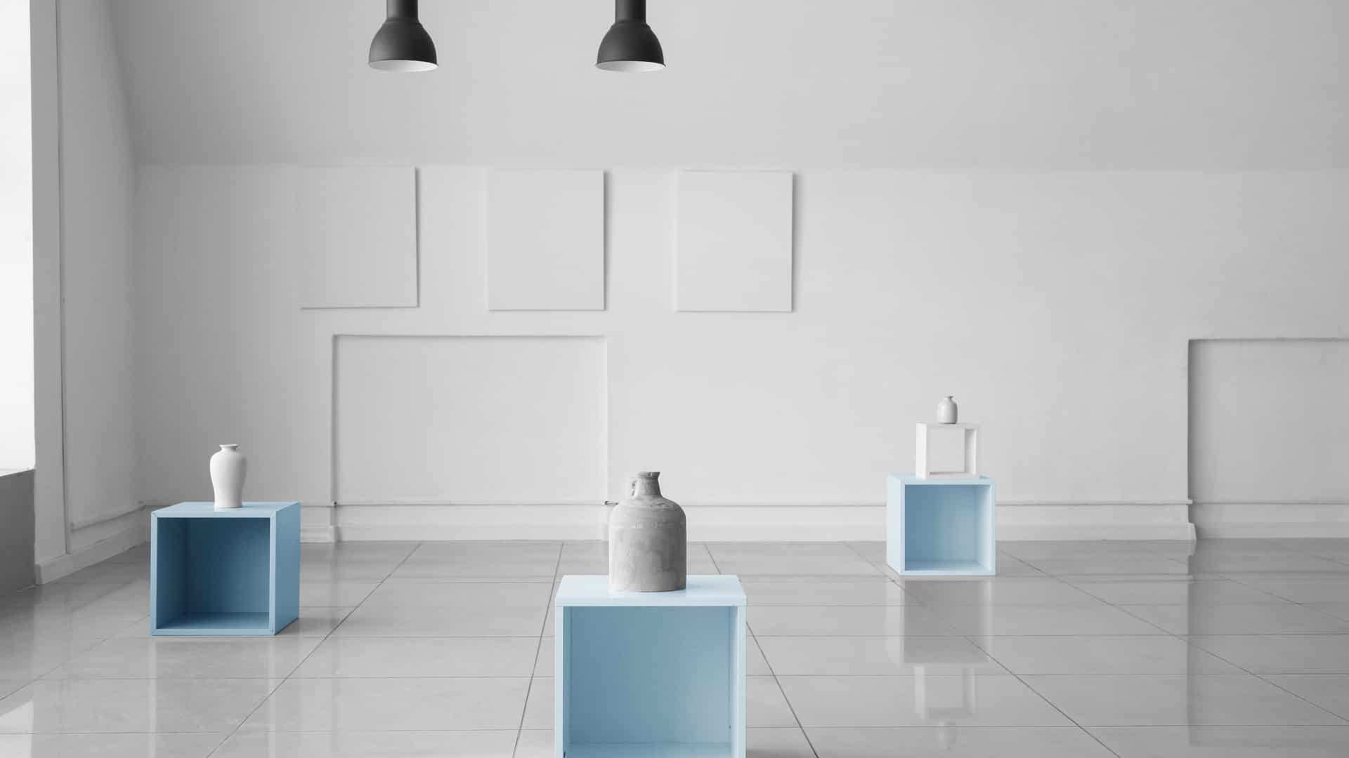 A sleek, modern "white cube" art gallery interior, reminiscent of the exhibition spaces at the Queensland Art Gallery and Gallery of Modern Art (QAGOMA). The room features a polished light colored floor, white walls, and three sky blue cube pedestals, each displaying a simple ceramic vase. Three rectangular white canvases hang blankly on the wall, lit by three black ceiling spotlights.