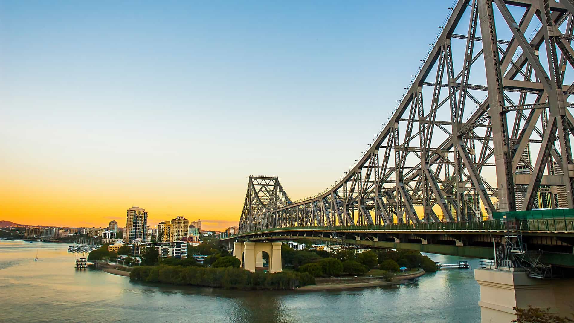 A scenic, wide angle photo of the Story Bridge in Brisbane, Australia, at dusk. The bridge's steel frame is illuminated with lights, creating a bright outline against the darkening sky. The image is taken from a low angle, showing the bridge from below as it spans across the Brisbane River. The city skyline is visible in the background, with the lights of the buildings starting to glow. The calm water of the river reflects the lights of the bridge and the city.