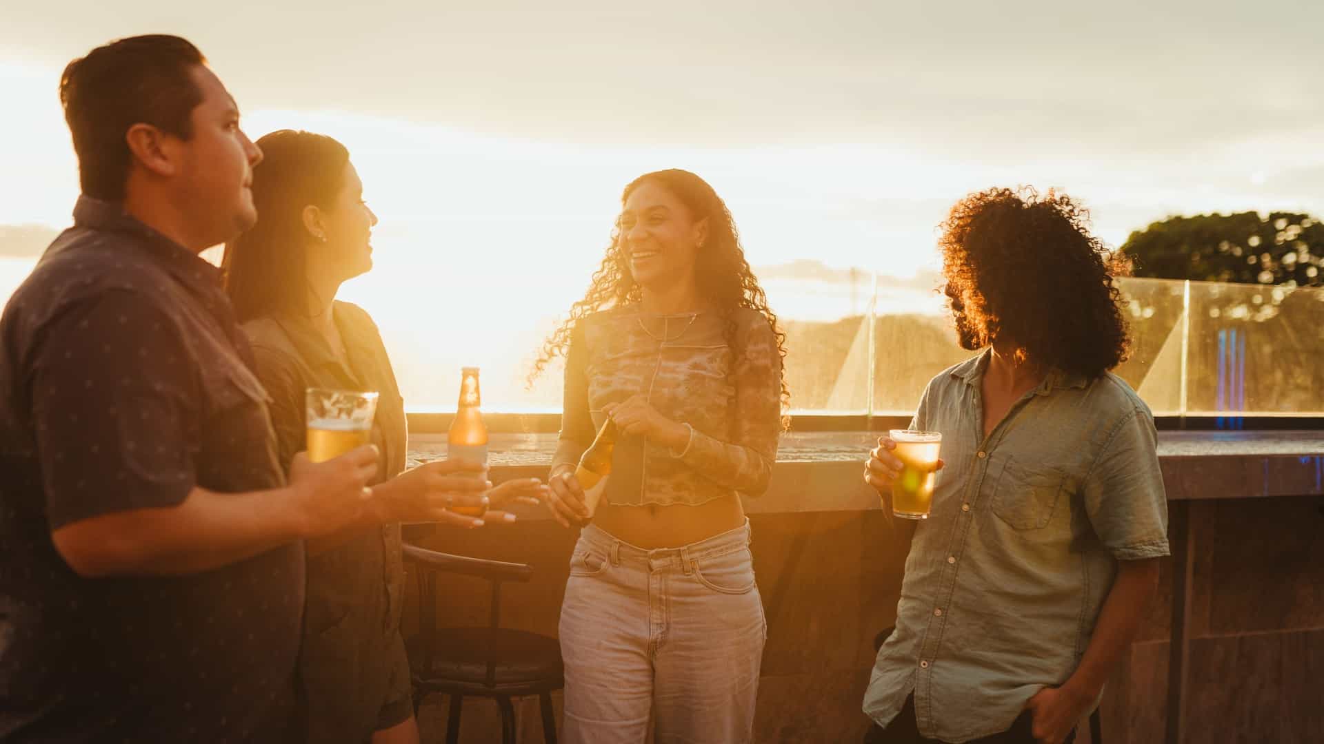 A group of young friends is enjoying Brisbane's sunset drinks at a rooftop bar, laughing and talking while holding beers, creating a vibrant scene of social interaction and leisure.