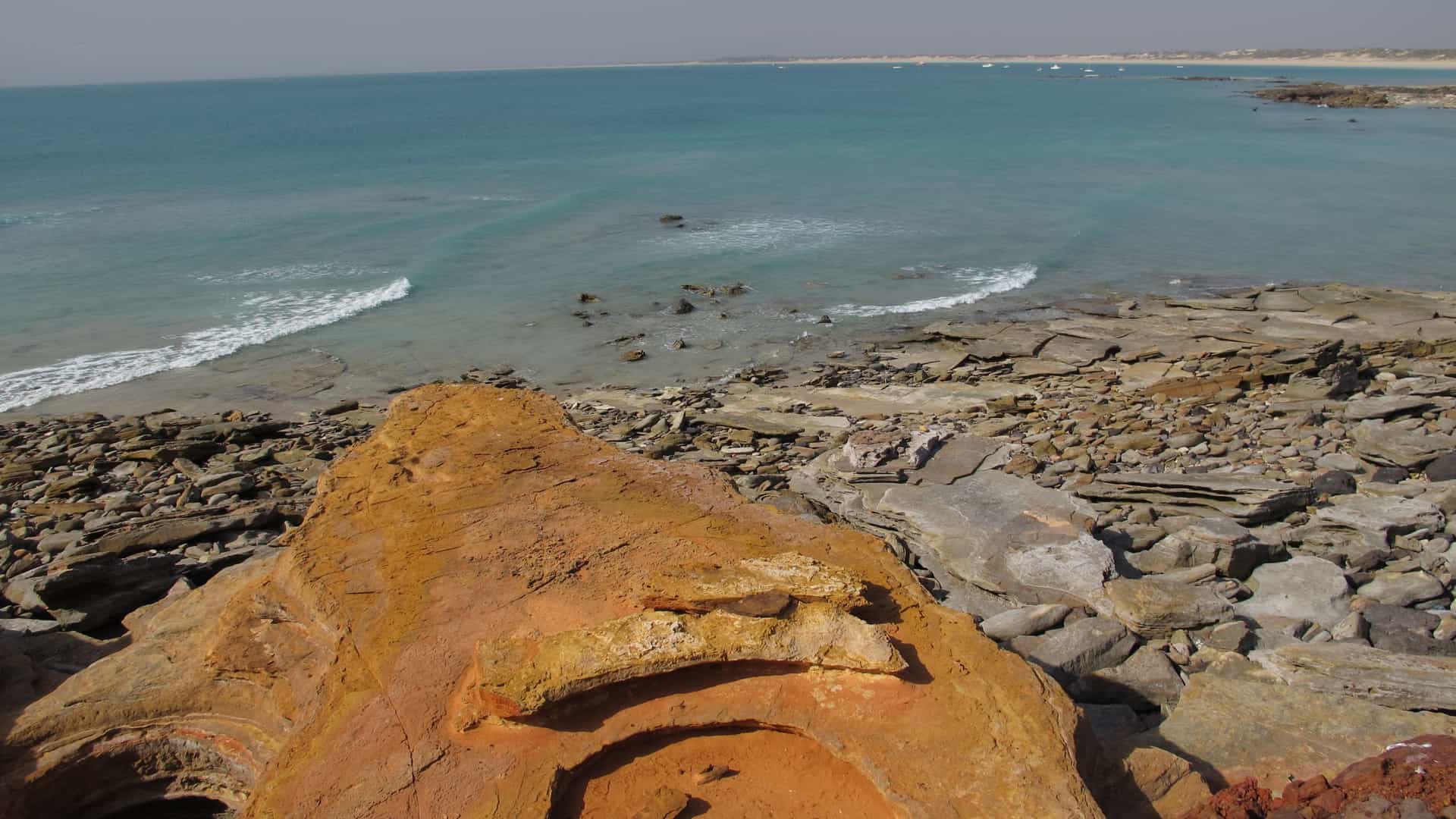 A scenic, horizontal photo of Gantheaume Point in Broome, Western Australia. The image is dominated by the vibrant colors of the landscape. On the left, there is a body of bright turquoise ocean water. On the right, the water meets a shoreline of white sand that curves toward the center of the frame. In the center and background, a large, dramatic promontory of deep red, rugged cliffs juts into the ocean. The cliffs have a layered, rocky texture and are a strong visual contrast to the blue water and white sand. The sky above is a clear, light blue with a few scattered white clouds.