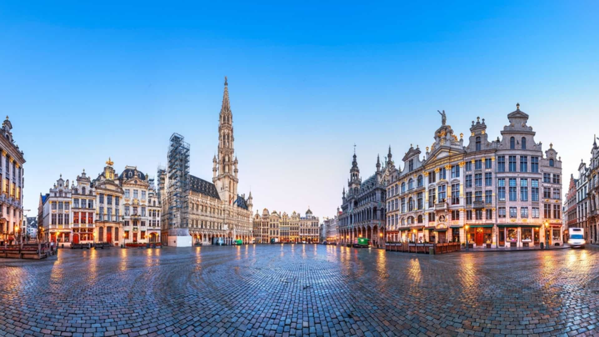 A scenic, horizontal photo of the Grand Place in Brussels, Belgium, illuminated at night. The central focus is the Brussels Town Hall, a large, ornate Gothic building with a towering, asymmetrical spire. The facade of the Town Hall is highly detailed with numerous statues and spires, and it is brightly lit, making it stand out against the dark sky. The square is cobblestone, and in the foreground, there are people walking around. The other buildings surrounding the square are also illuminated, showcasing their ornate Baroque architecture, with gilded details catching the light. The overall scene is lively and historic.