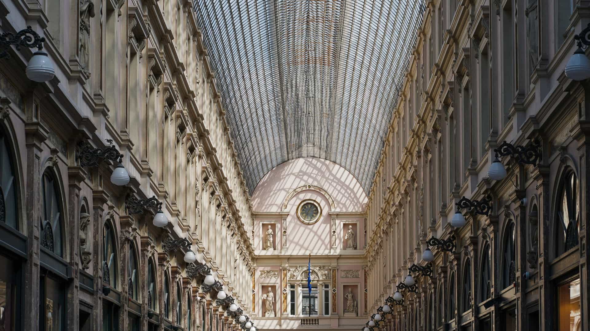The elegant, ornate glass and iron roof of the Galeries Royales Saint Hubert in Brussels, Belgium, spans the historic shopping arcade with intricate architectural details visible from the ground floor.