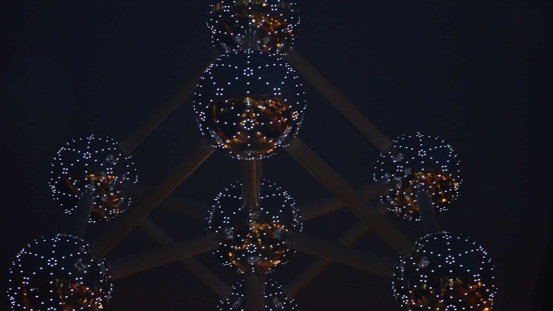 A dramatic night photo of the Atomium in Brussels, Belgium, an iconic structure representing an iron crystal magnified 165 billion times. The nine spheres and connecting tubes are brightly illuminated by thousands of lights against a dark night sky.