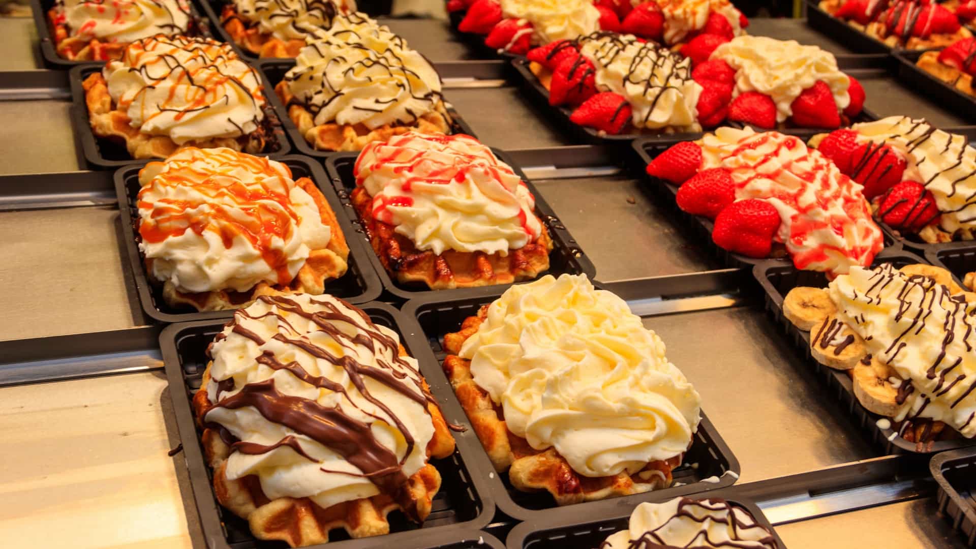 A close up display of several Liege style Belgian waffles with a variety of colorful and sweet toppings, including fresh fruits like strawberries and kiwi, whipped cream, melted chocolate, and brightly colored glazes, displayed for sale in a food stall.
