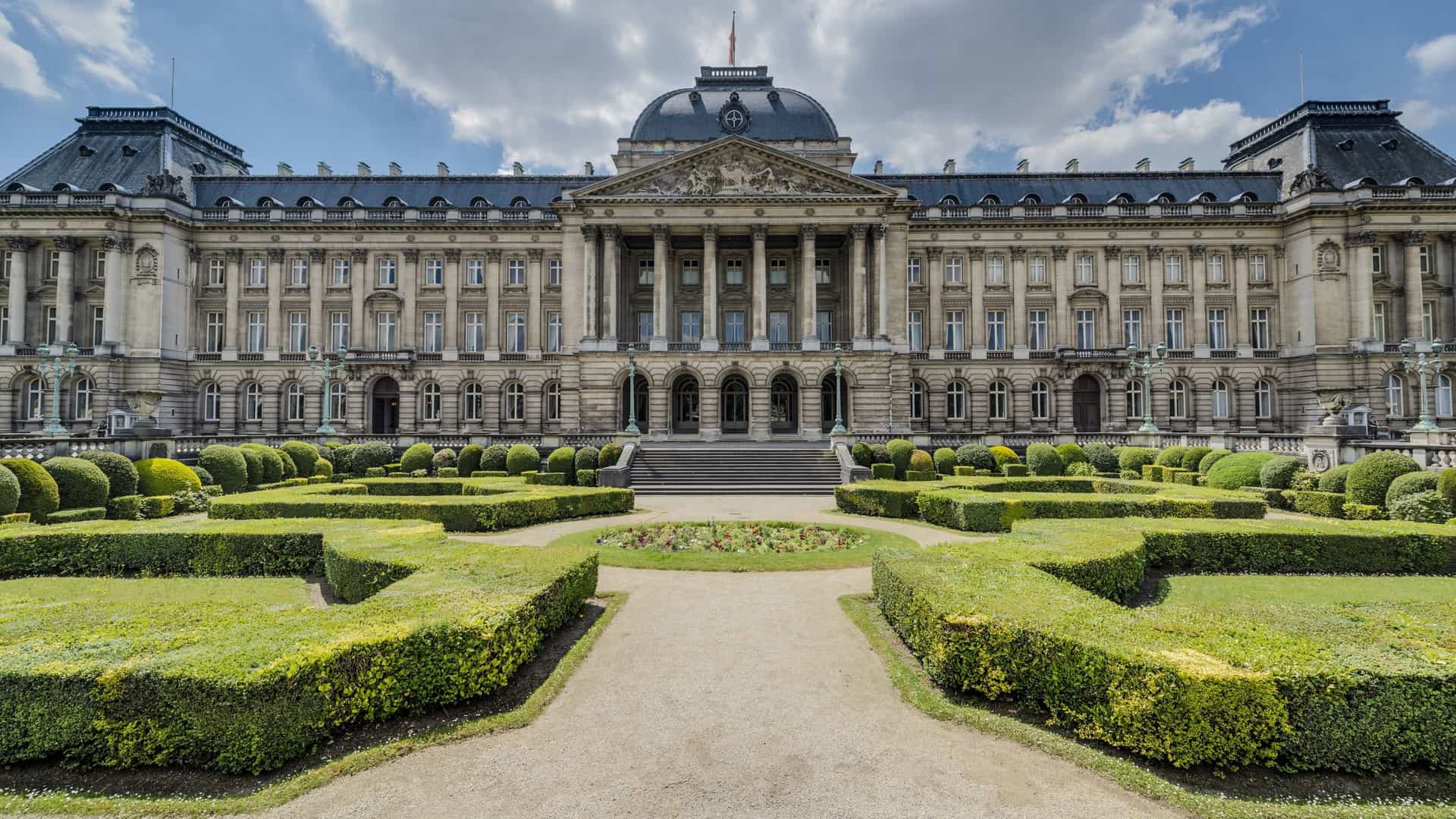 A scenic, wide angle photo of the Royal Palace of Brussels during the daytime. The palace is a large, symmetrical neoclassical building with a central, prominent facade. The building has a detailed white or light colored stone facade with many windows, pillars, and a central triangular pediment. In front of the palace is a large open square, the Place des Palais. The sky is bright and blue, and there are trees and a park visible in the foreground and on either side of the palace, adding a touch of nature to the formal, urban scene.