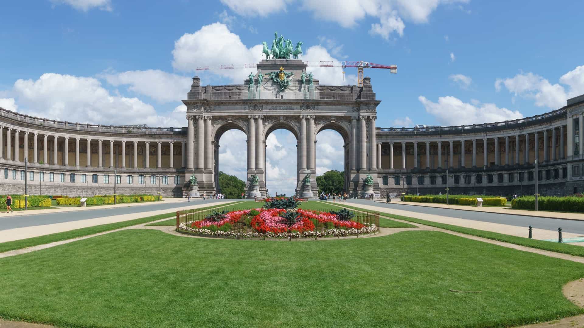 The Parc du Cinquantenaire's triumphal arch in Brussels, Belgium. The large, three arched stone monument is topped by a bronze sculpture of a quadriga, a four horse chariot with a charioteer. The arch is surrounded by a large public park with green lawns and trees.