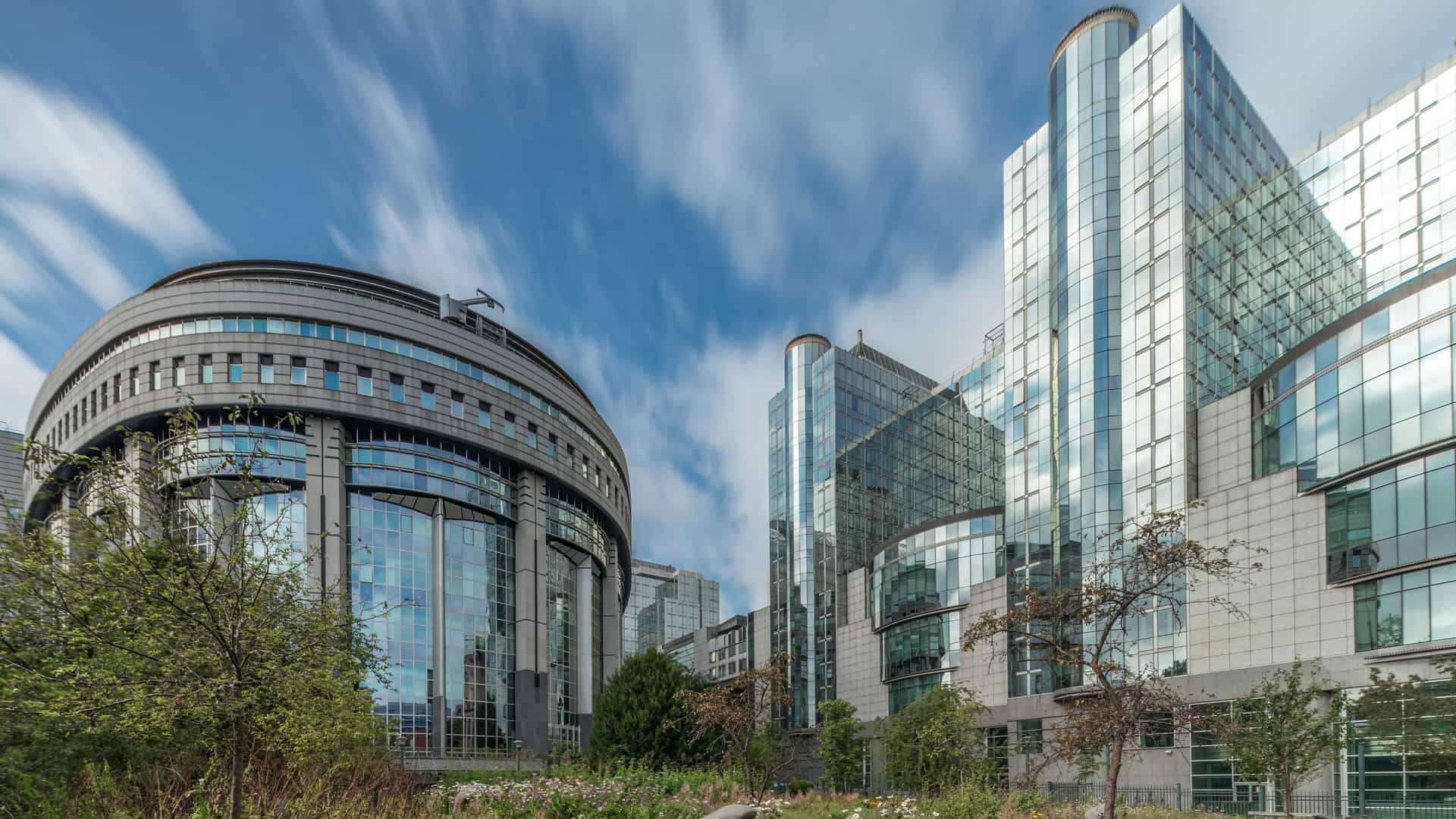 A timelapse of the European Quarter in Brussels, with modern glass facade buildings, including the European Parliament and Commission, visible under a dramatic cloudy sky. In the foreground is a park with green lawns and trees.