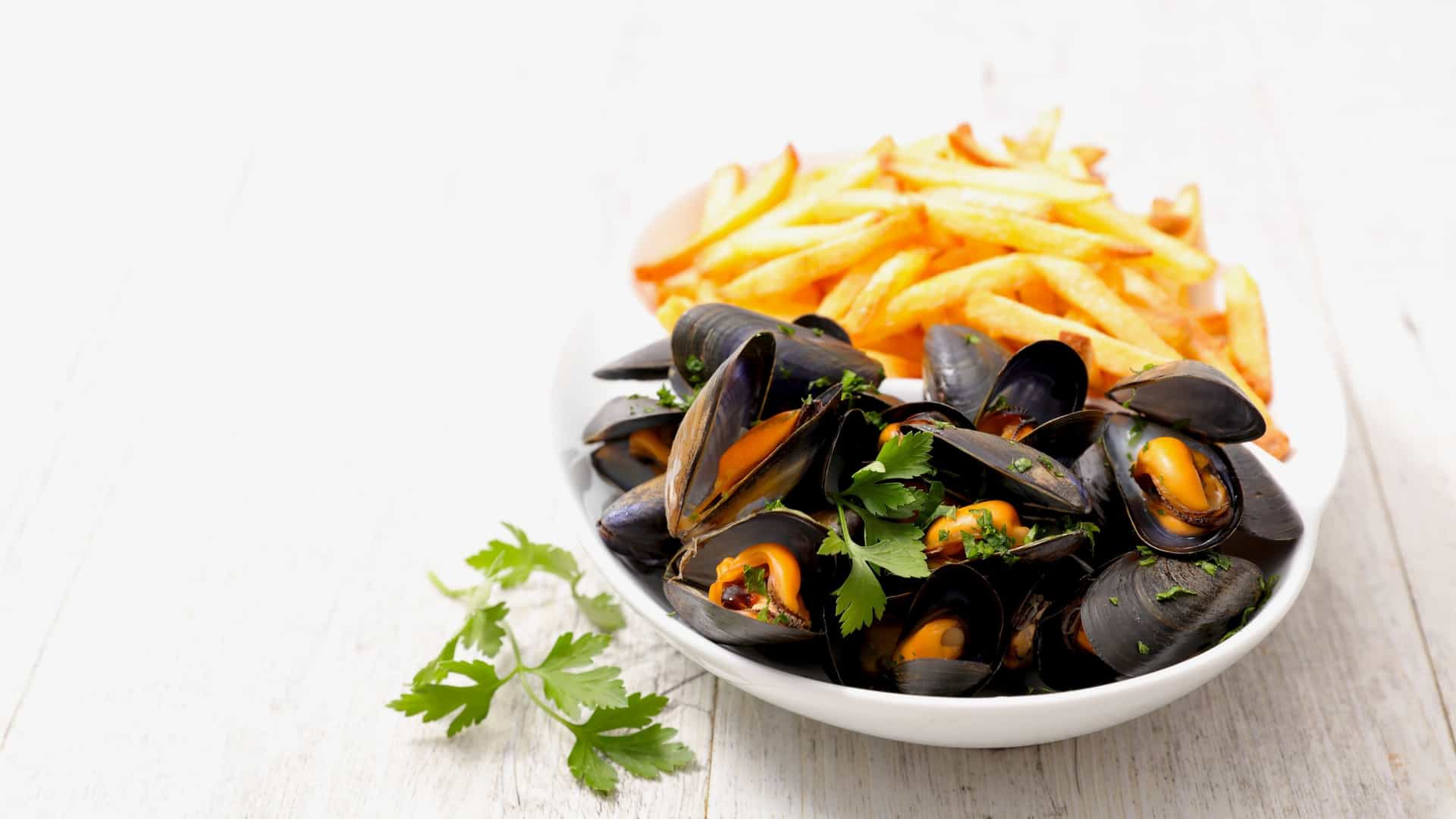 A traditional Belgian and French dish of Moules Frites (mussels and fries) served on a table, featuring a large metal pot filled with steamed mussels alongside a conical paper holder overflowing with golden, crispy French fries.