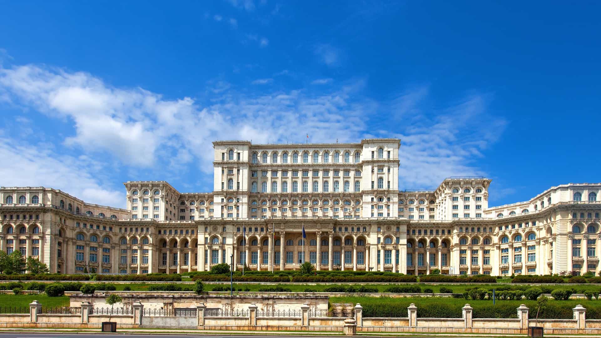 The imposing facade of the Palace of the Parliament in Bucharest, Romania. The massive, multi story building features neoclassical architecture with numerous windows, columns, and an ornate exterior. It is a dominant landmark on the Bucharest skyline, visible under a bright sky.