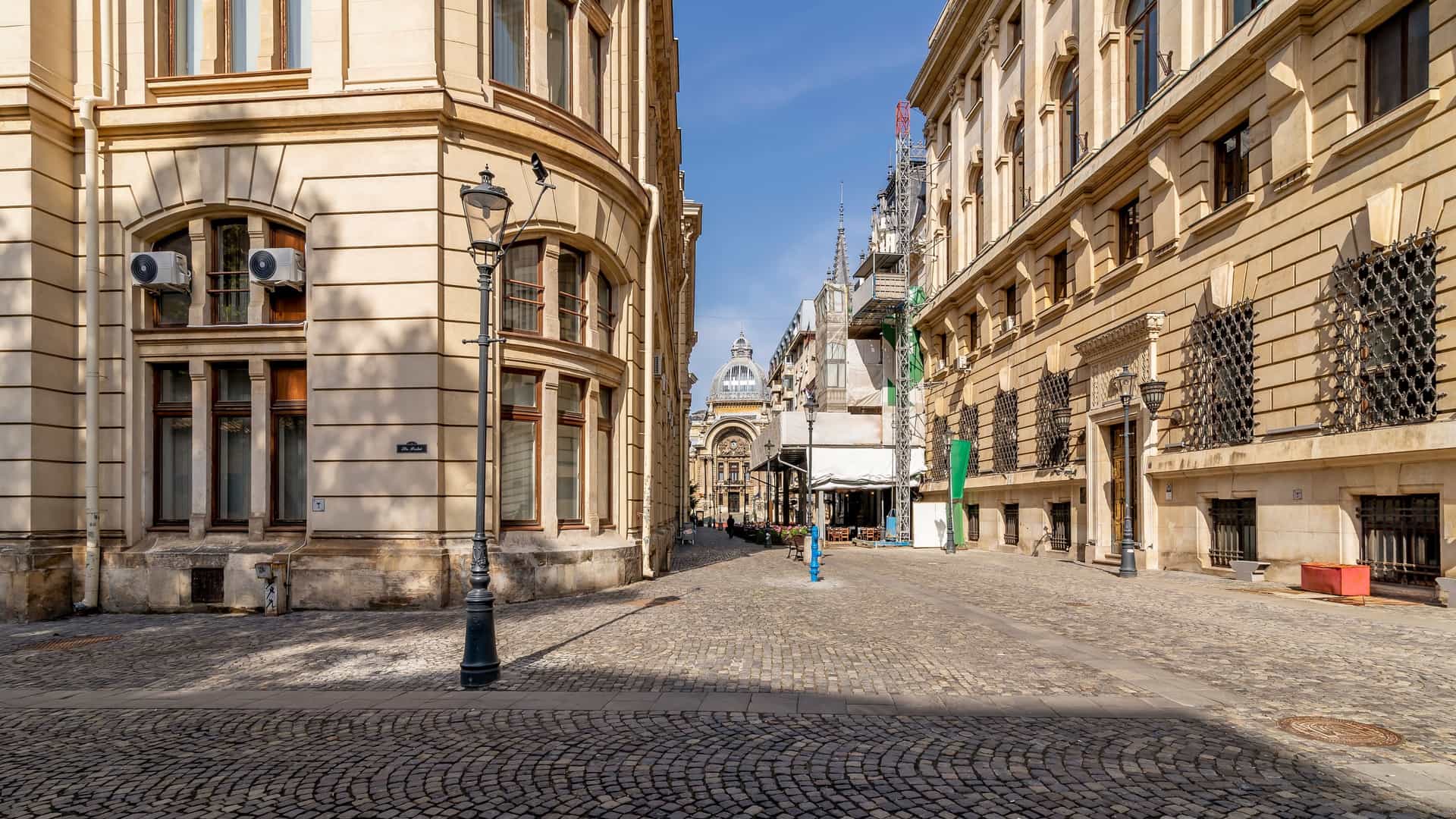 A view of the intersection of Stavropoleos and Lipscani streets in the Old Town of Bucharest, Romania, showing a narrow cobblestone pedestrian street lined with historic buildings featuring classical architecture and storefronts on a sunny day. The buildings have varying colors, and a number of people are walking down the street.