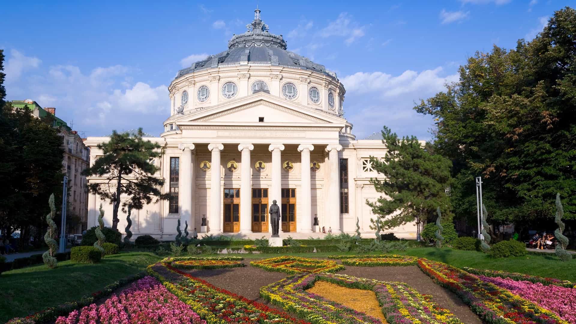 A wide shot of Piața George Enescu in Bucharest, Romania, featuring the iconic dome and columned facade of the Romanian Athenaeum concert hall, with a statue visible in the square.
