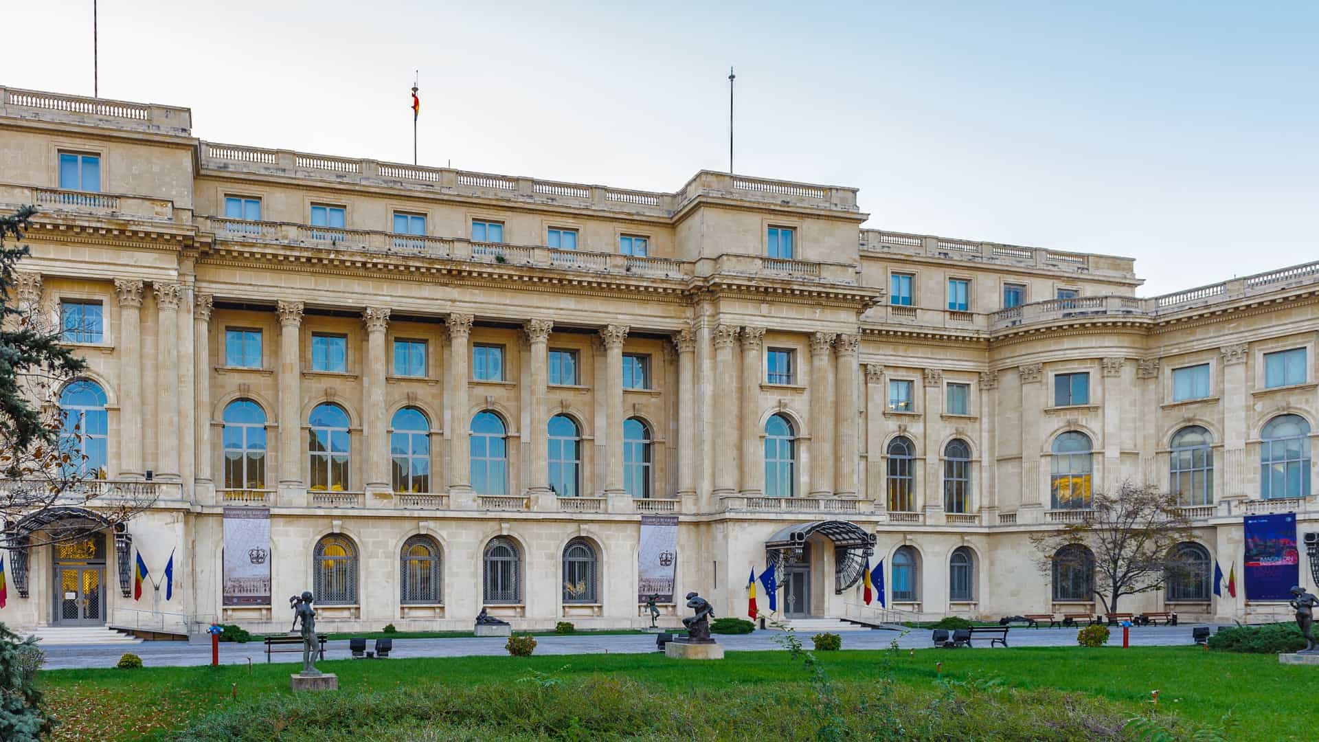 The main entrance of the National Museum of Art of Romania (Muzeul Național de Artă al României) in Bucharest, which is housed in the former Royal Palace. The neoclassical building features grand columns and a large, ornate arched doorway, set against the backdrop of Revolution Square.