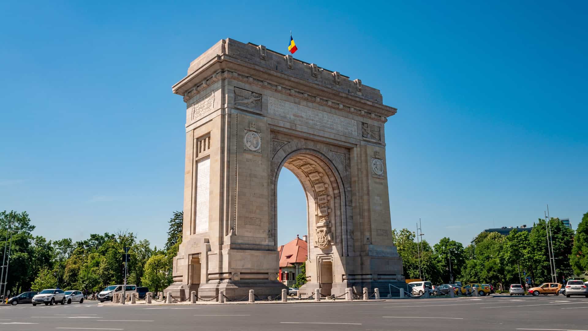 The Triumphal Arch (Arcul de Triumf) in Bucharest, Romania, stands majestically on Kiseleff Road in the northern part of the city. The monumental arch, built of Deva granite and modeled after the Arc de Triomphe in Paris, is captured against a clear blue sky on a sunny day, showcasing its neoclassical architecture and intricate sculptural details. It is a symbol commemorating Romania's victory in World War I and the Great Union of 1918.