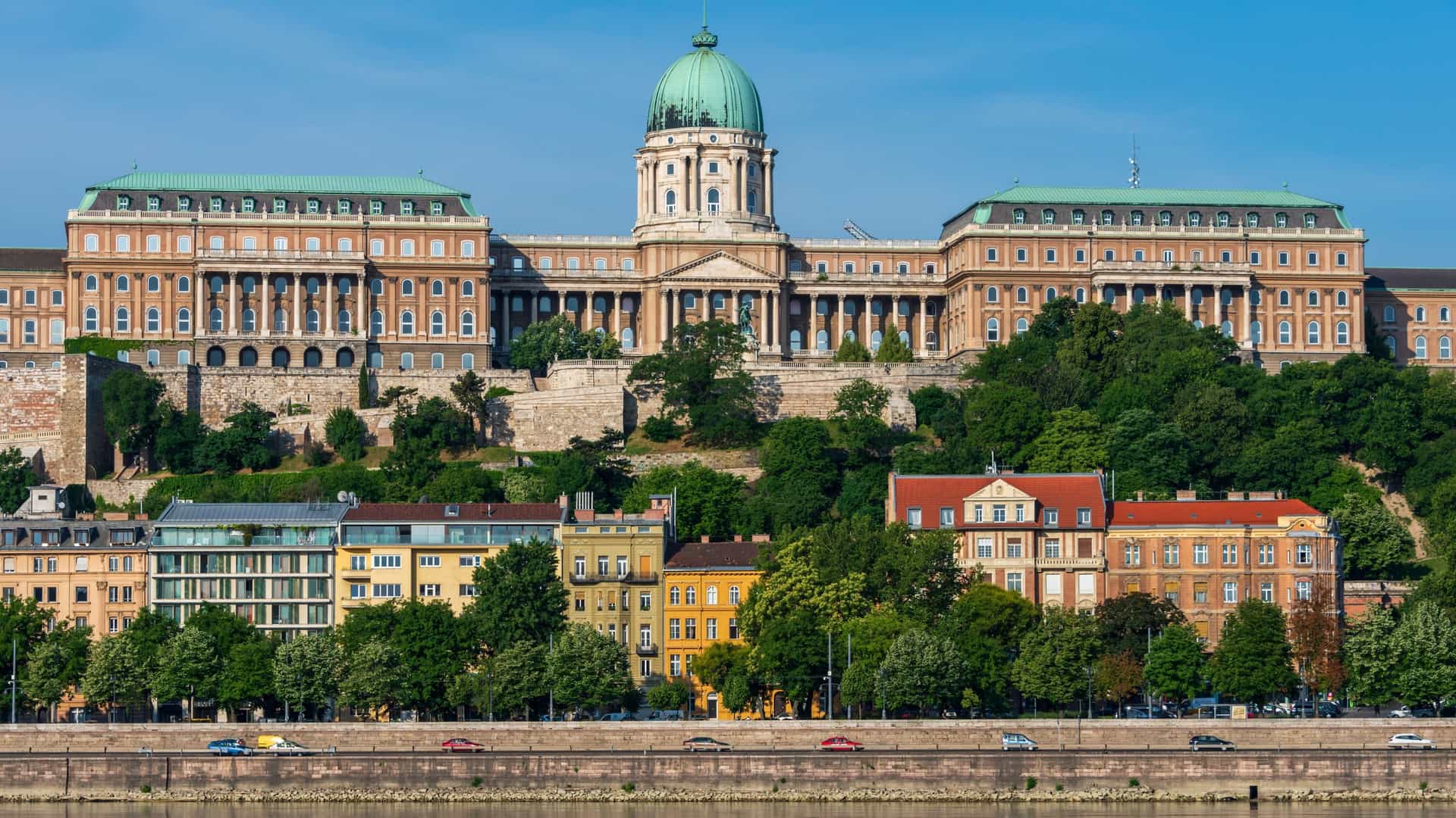 Panoramic daytime view of the Budapest skyline, featuring the historic Buda Castle and Royal Palace dominating Castle Hill in the foreground. The wide Danube River separates Buda from Pest, where the Chain Bridge, the Hungarian Parliament Building, and St. Stephen's Basilica are visible landmarks.