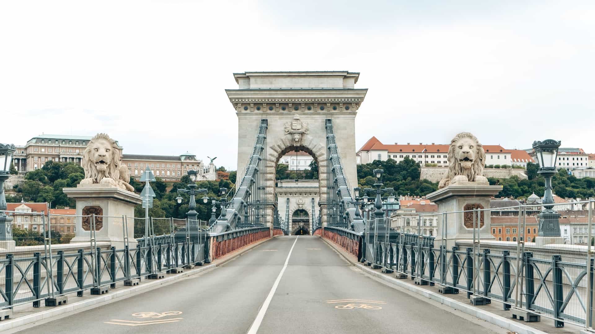 A scenic night view of the Széchenyi Chain Bridge spanning the Danube River in Budapest, Hungary. The historic suspension bridge is brightly lit, and its lights are reflected on the water's surface. The bridge connects the illuminated cities of Buda and Pest, with the Buda Castle visible in the background on the hill.