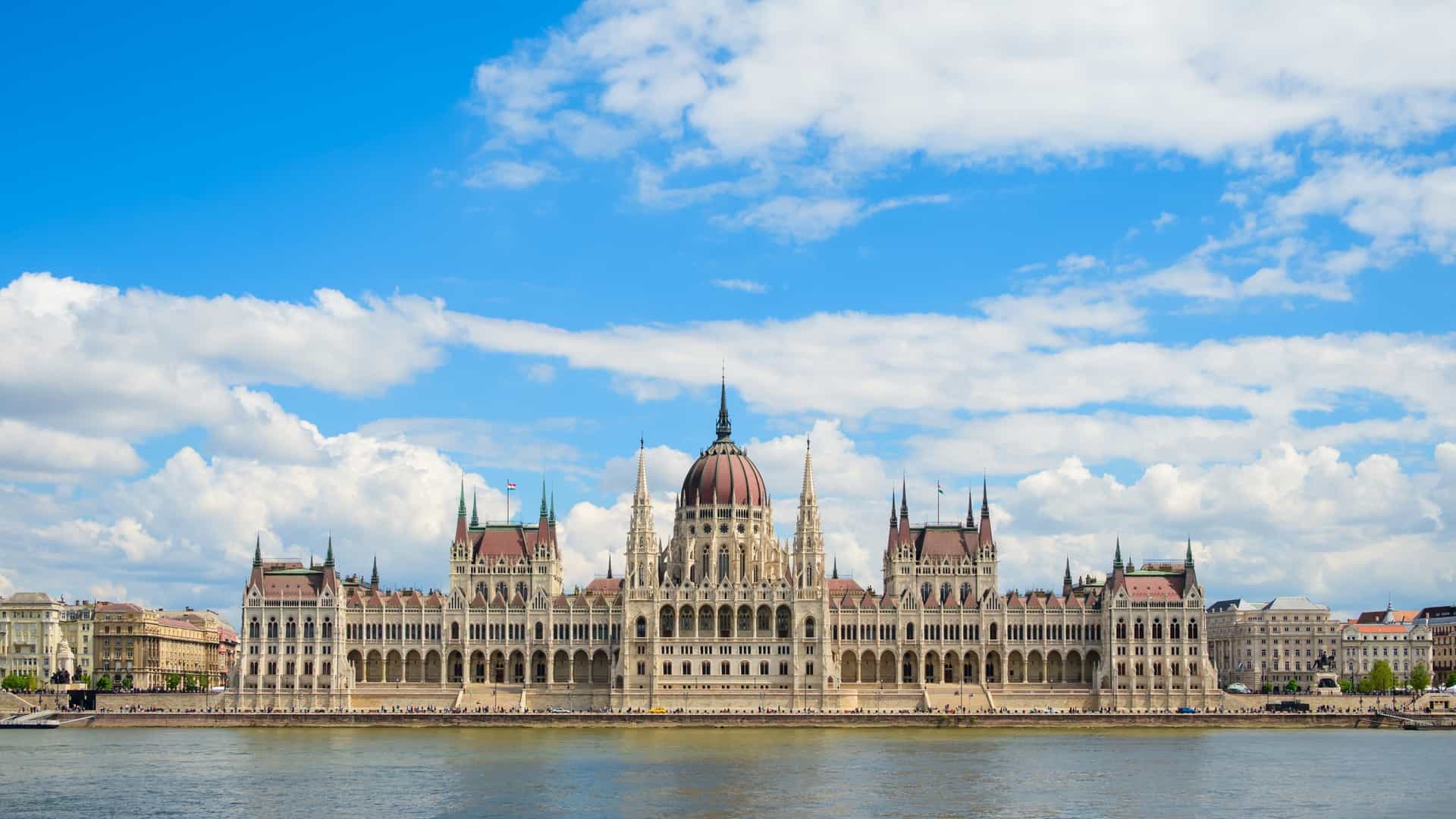 A daytime photo of the Hungarian Parliament Building in Budapest, Hungary. The neo Gothic building stands prominently along the Danube River under a bright blue sky.
