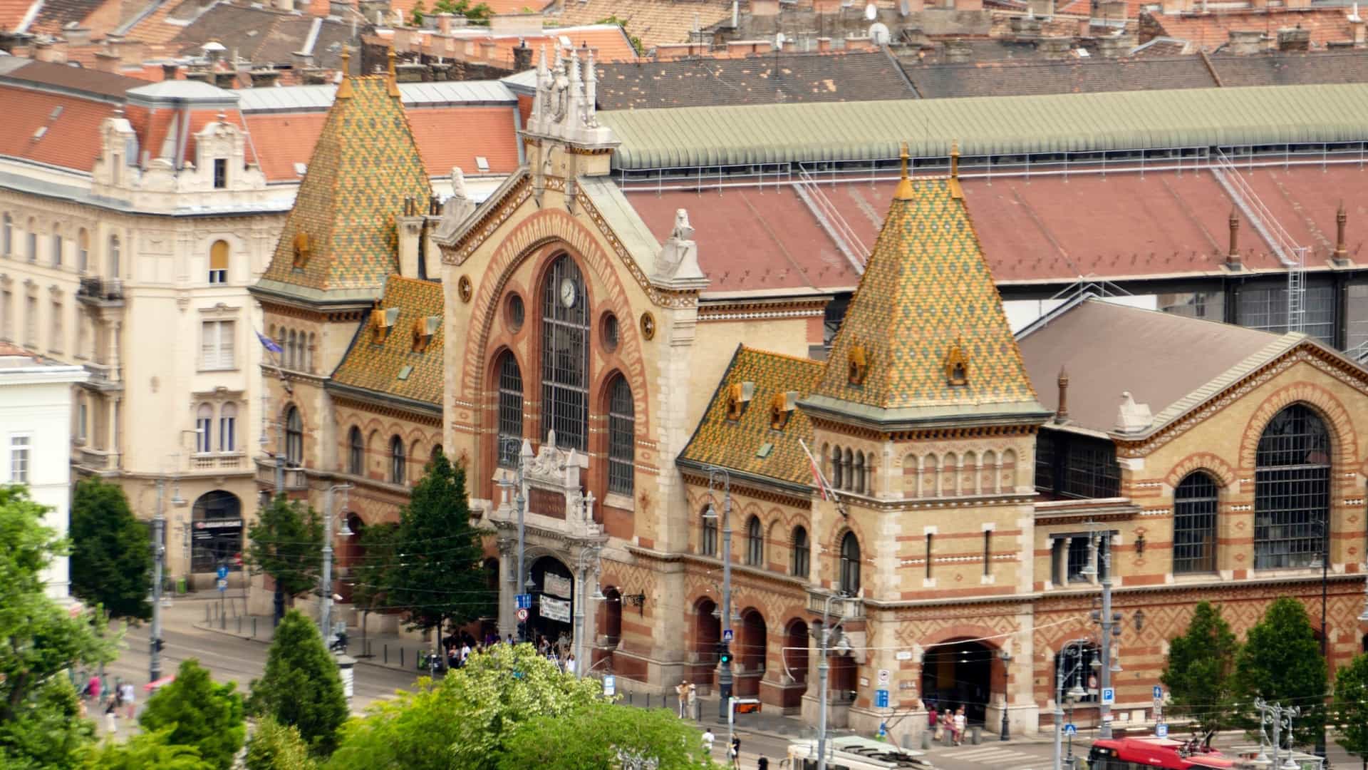 An exterior, wide angle view of the Central Market Hall (Nagyvásárcsarnok) in Budapest, Hungary. This historical, neo Gothic building features large brick walls, stone archways, and a distinctive roof covered with colorful Zsolnay ceramic tiles. The photo is taken on a sunny day, showcasing the facade and the building's grandeur.