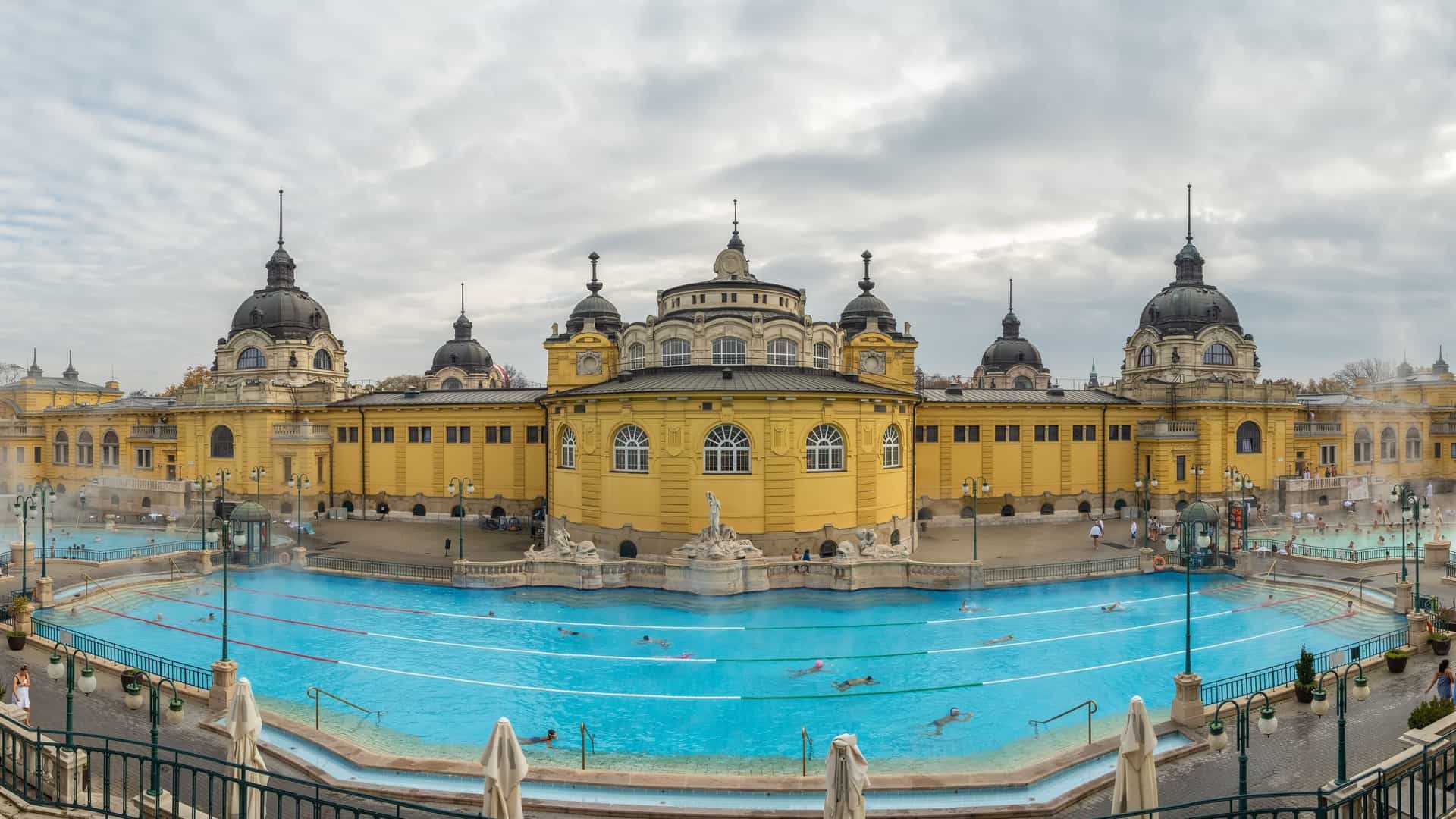 A panoramic photo of the historical Széchenyi Baths in Budapest, Hungary, in winter. The Neo Baroque building is seen with steam rising from the outdoor thermal pools, where people are bathing in the cold weather.