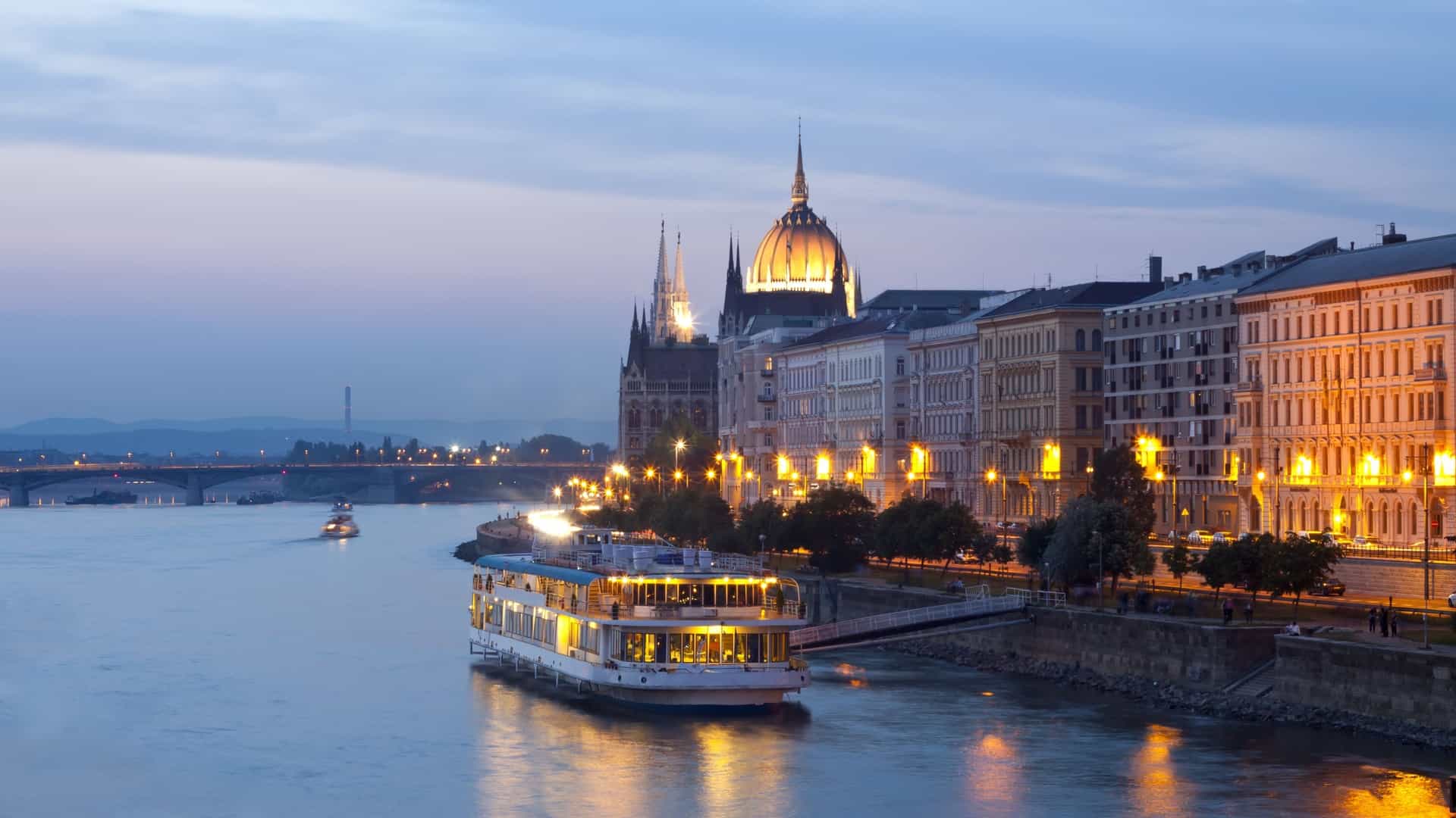 A night photo of Budapest with a small illuminated cruise boat on the Danube River, showing the city's landmarks lit up in the background.