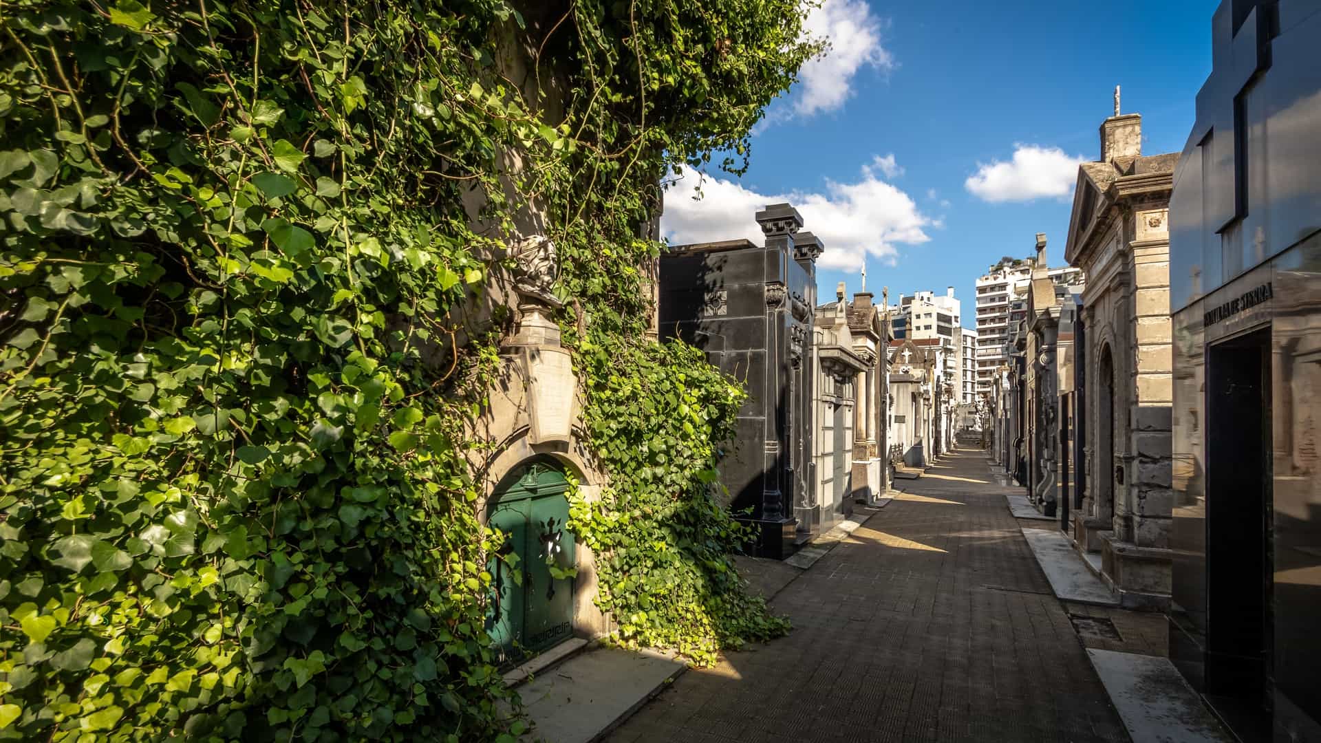 An alleyway within the Recoleta Cemetery (Cementerio de la Recoleta) in Buenos Aires, Argentina. The above ground mausoleums, built in various opulent architectural styles like Neo Gothic and Art Nouveau, line the path like miniature city blocks, showcasing detailed marble statues and ornate ironwork gates. The grand, yet often weathered, tombs give the cemetery the appearance of a lavish city of the dead.