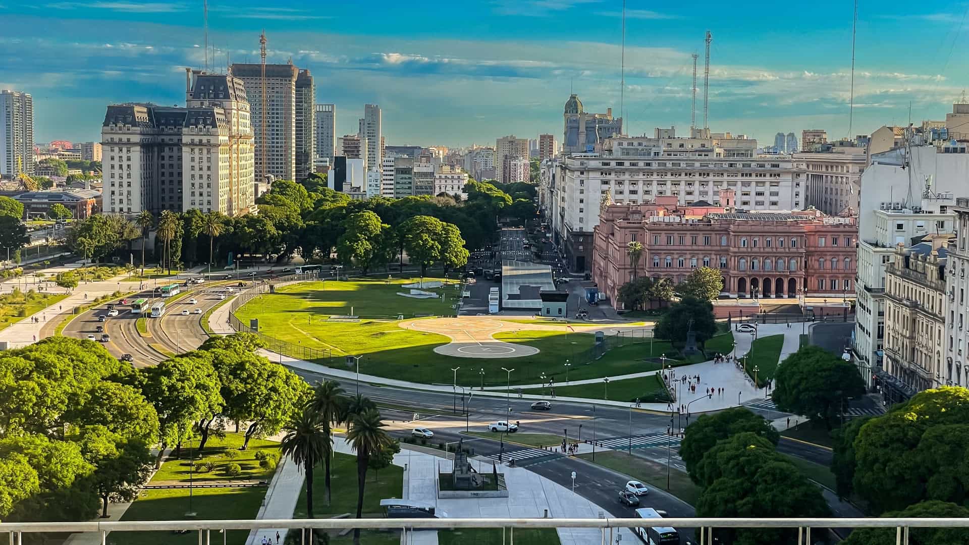 A stunning aerial view of Plaza de Mayo in Buenos Aires, Argentina, featuring the iconic Casa Rosada (the presidential palace, visible with its distinct pink color) at one end. The historic main square is surrounded by other major landmarks, including the Buenos Aires Cabildo (a white colonial building), and the Metropolitan Cathedral. The photograph captures the symmetrical design of the plaza, with the Pirámide de Mayo at its center, surrounded by trees and urban traffic, all under a clear sky.