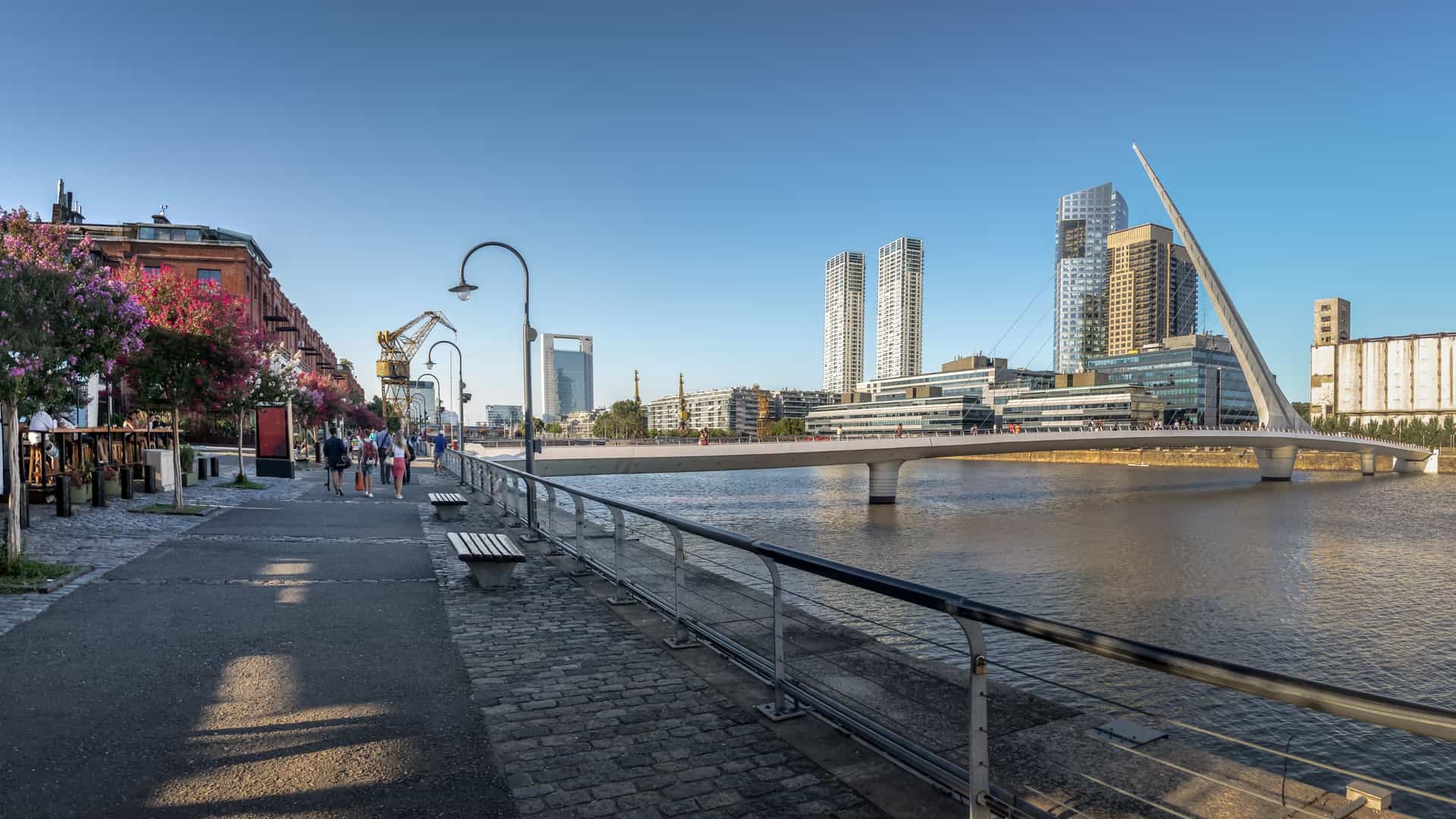 A panoramic photo of the Puerto Madero district in Buenos Aires, Argentina, featuring the Puente de la Mujer (Woman's Bridge), a modern pedestrian bridge with a distinctive, asymmetrical mast, spanning a dock surrounded by contemporary buildings.