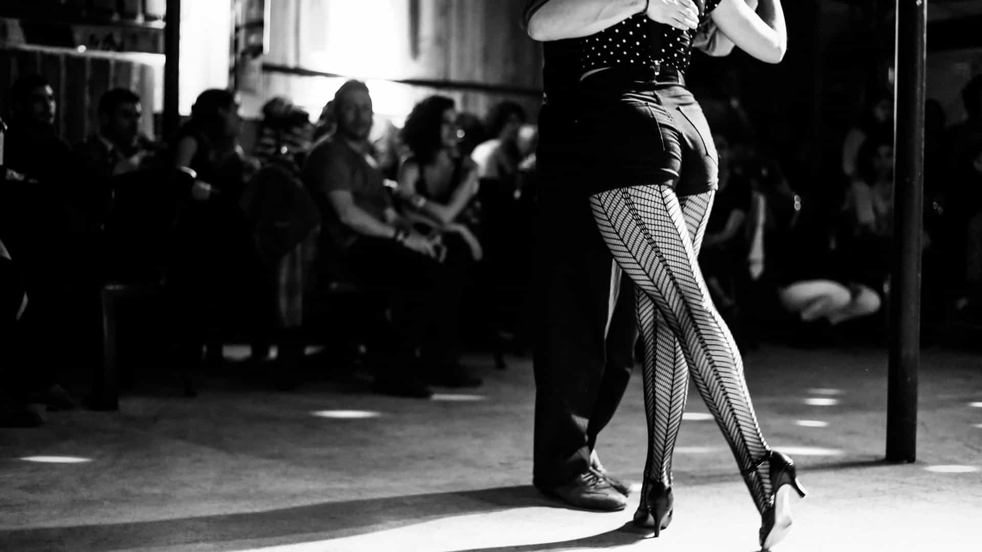 A passionate and close up view of a couple dancing the Tango in a milonga (tango dance hall) in Buenos Aires. The dancers are captured mid movement in a low key, dramatic light that highlights the intensity of the embrace. The male dancer is wearing a dark suit, and the female dancer is wearing a flowing, dark skirt with a slit, emphasizing the intimate and focused atmosphere of the social dance.